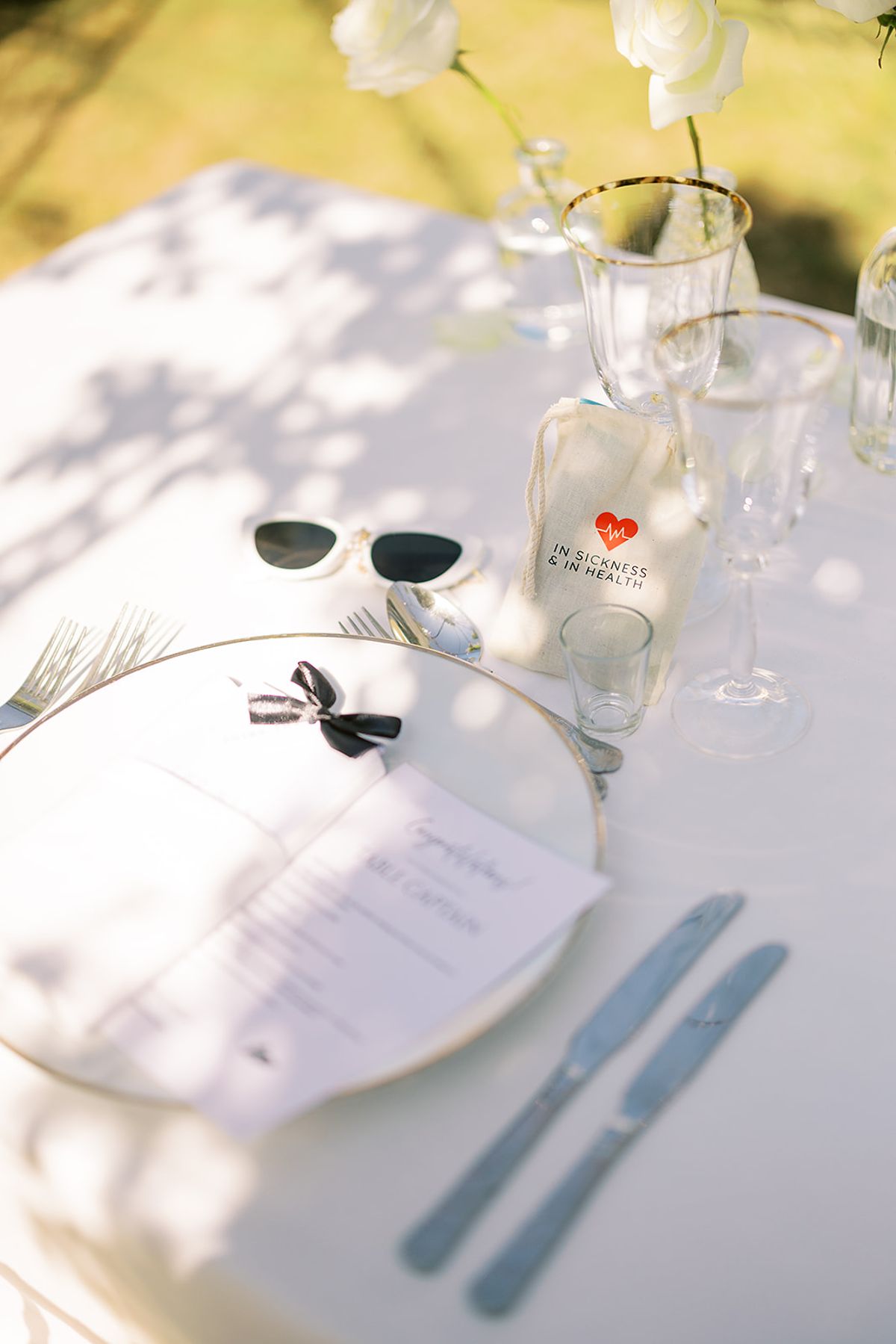 A white-clothed table set with a clear charger plate, printed menu, cutlery, glasses, a small bottle and a pair of sunglasses in dappled light.