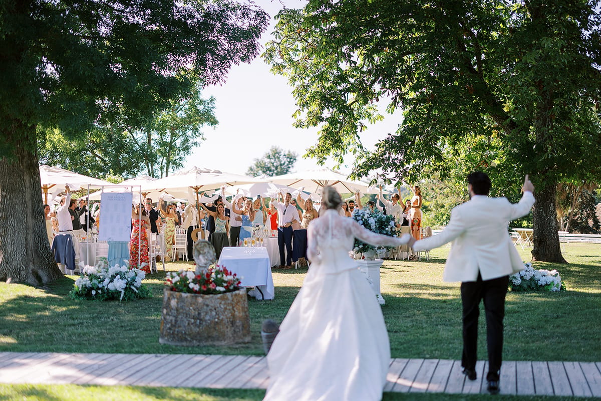The couple, seen from behind, walk hand in hand towards cheering guests with raised arms beneath white parasols on a lawn.