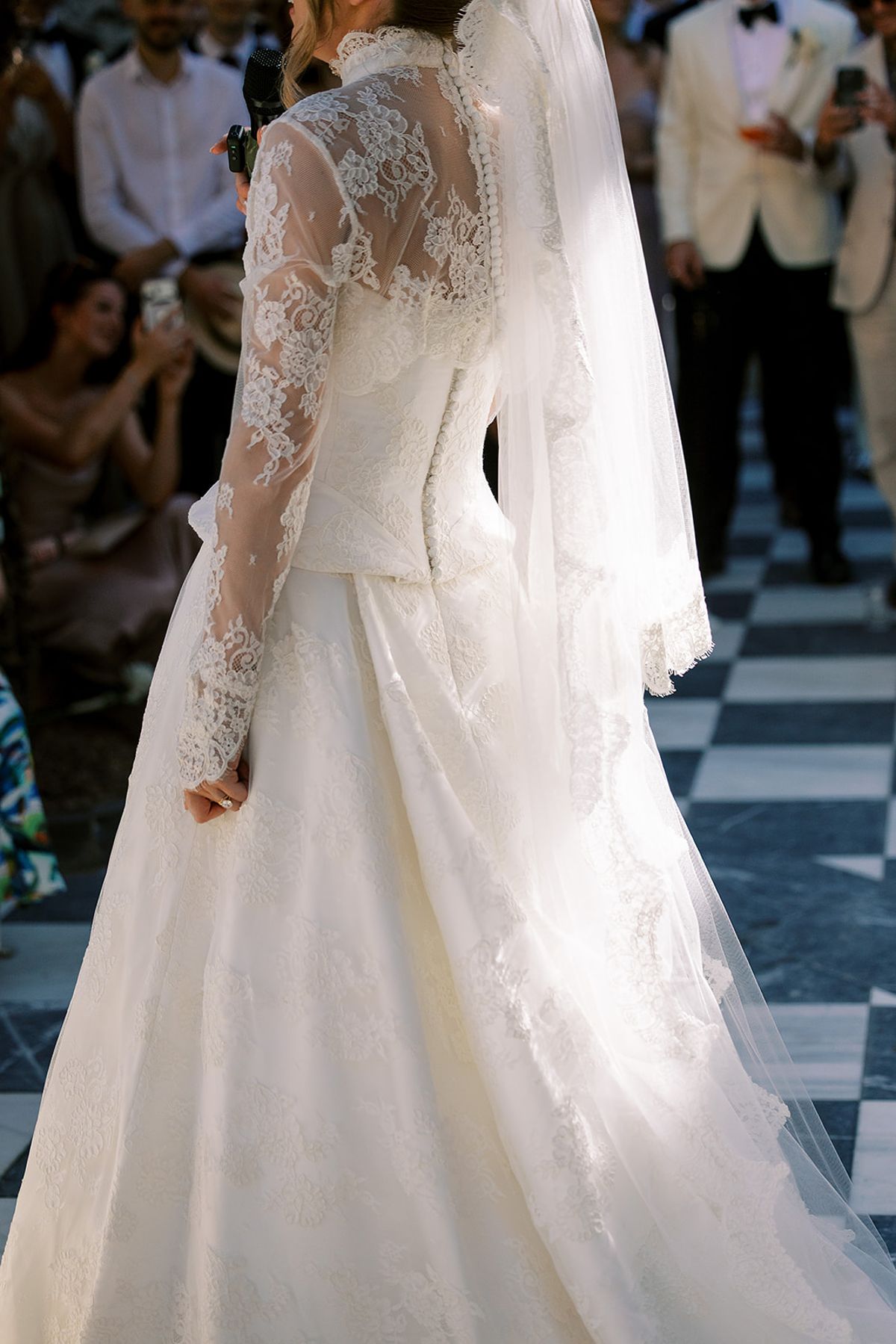 The bride, seen from behind in a long-sleeved lace gown and veil, holds a microphone as guests watch on a chequered floor.