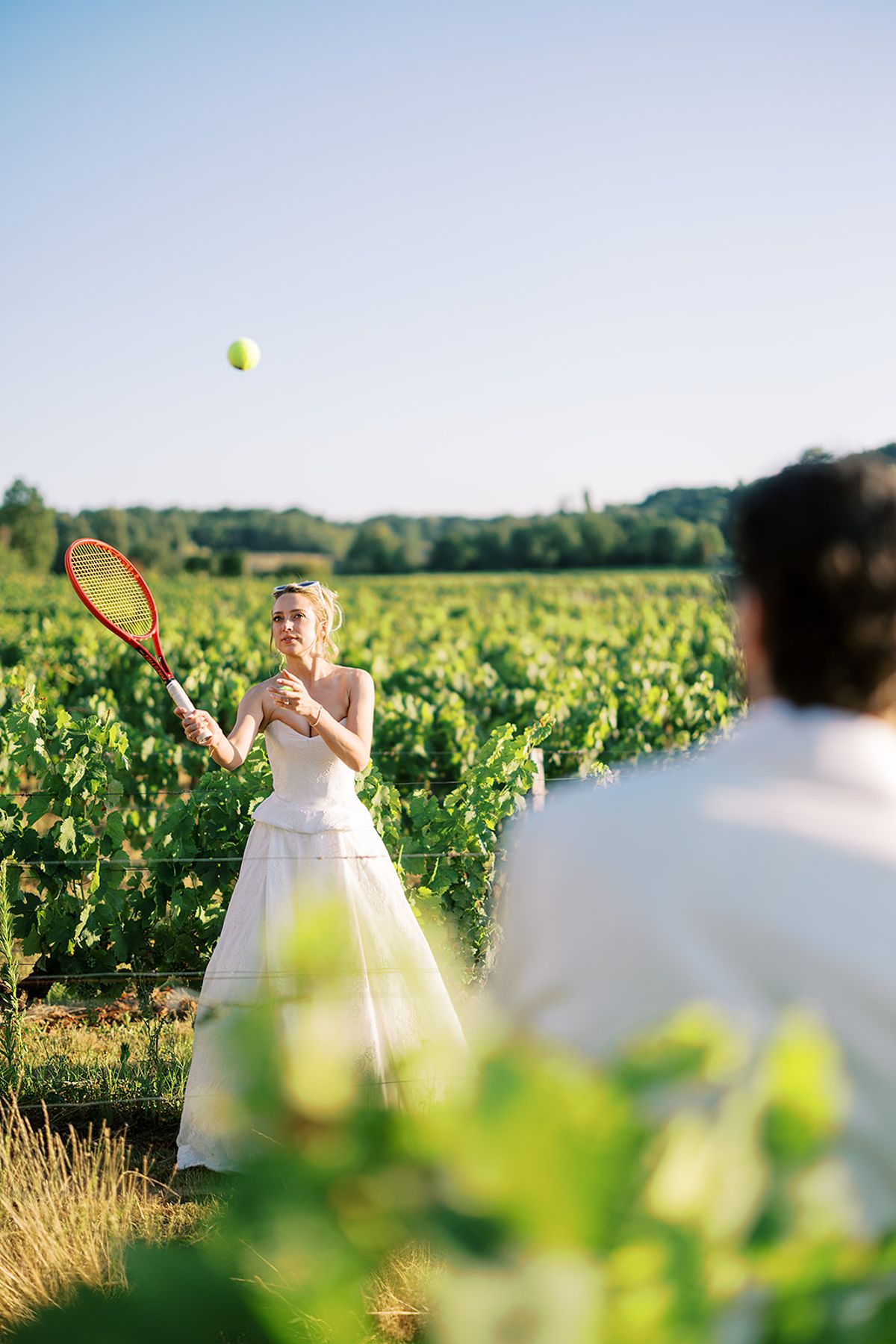 The bride in a white gown holds a tennis racket and tosses a ball in a vineyard, with the groom blurred in the foreground.