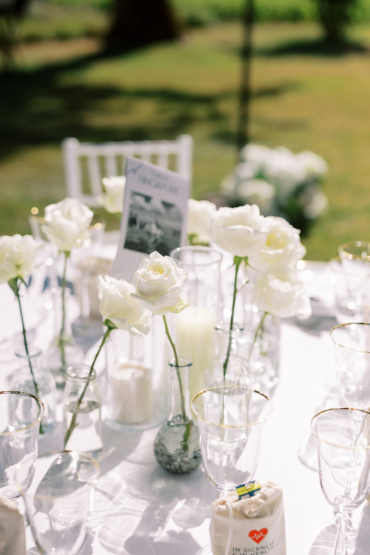 A round table set with white roses in slim vases, candles, glassware and a small printed card.