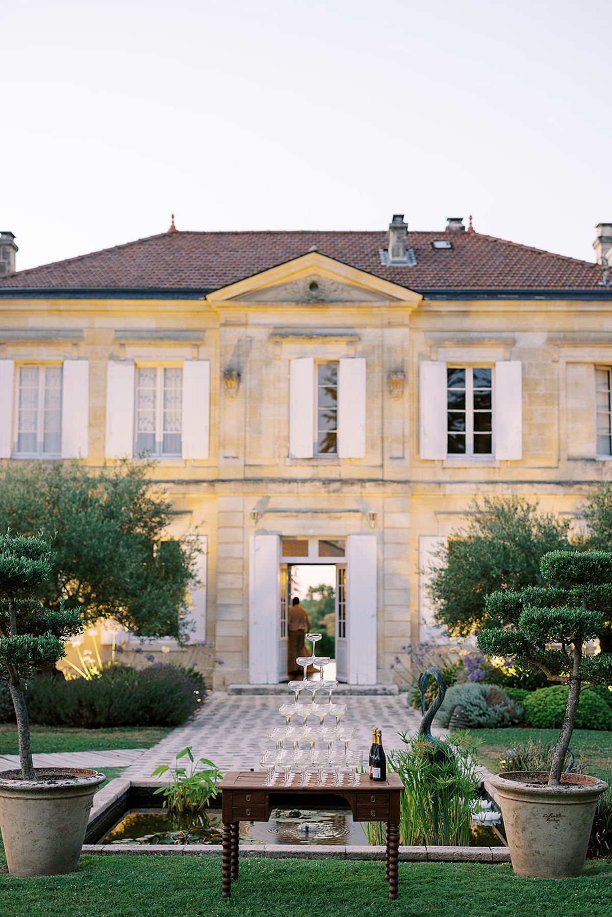 A person stands in the open doorway of a stone château; a champagne glass tower and bottle sit on a table in the garden.