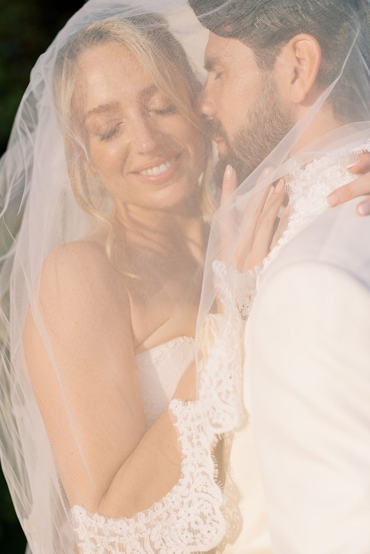 The groom kisses the smiling bride's cheek as they embrace, both softly covered by her sheer veil with a lace edge.