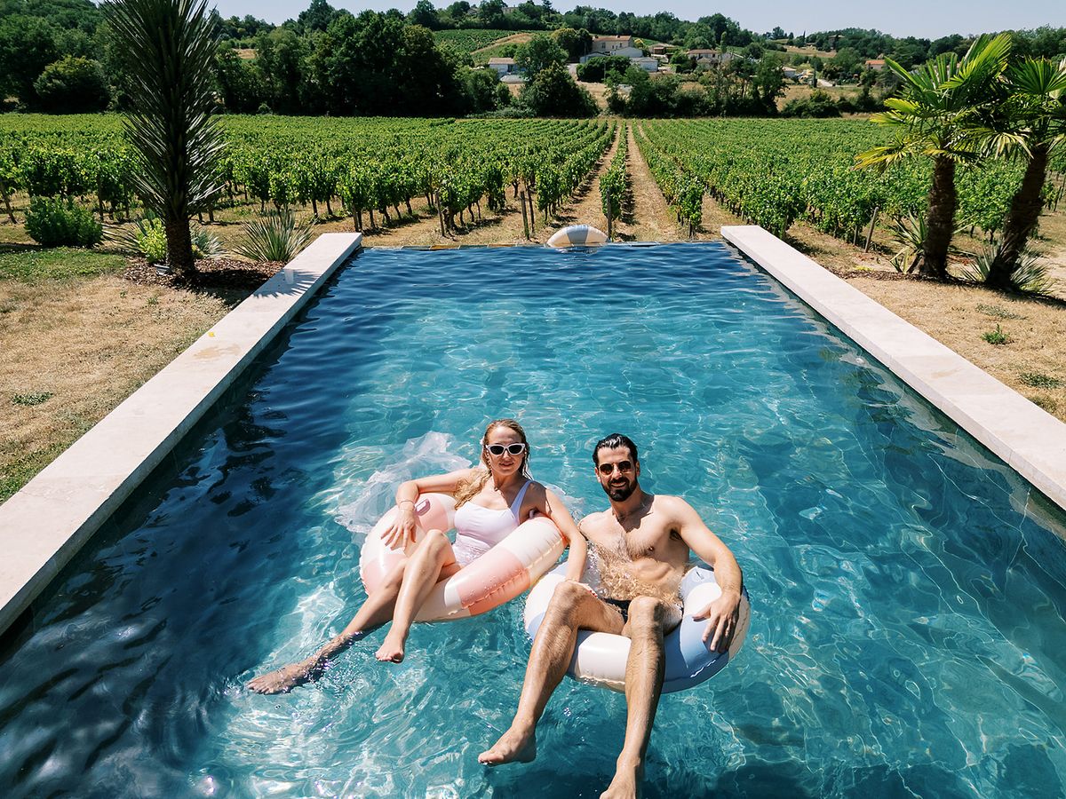The couple float side by side on inflatable rings in a clear blue pool, with rows of vines stretching behind them.