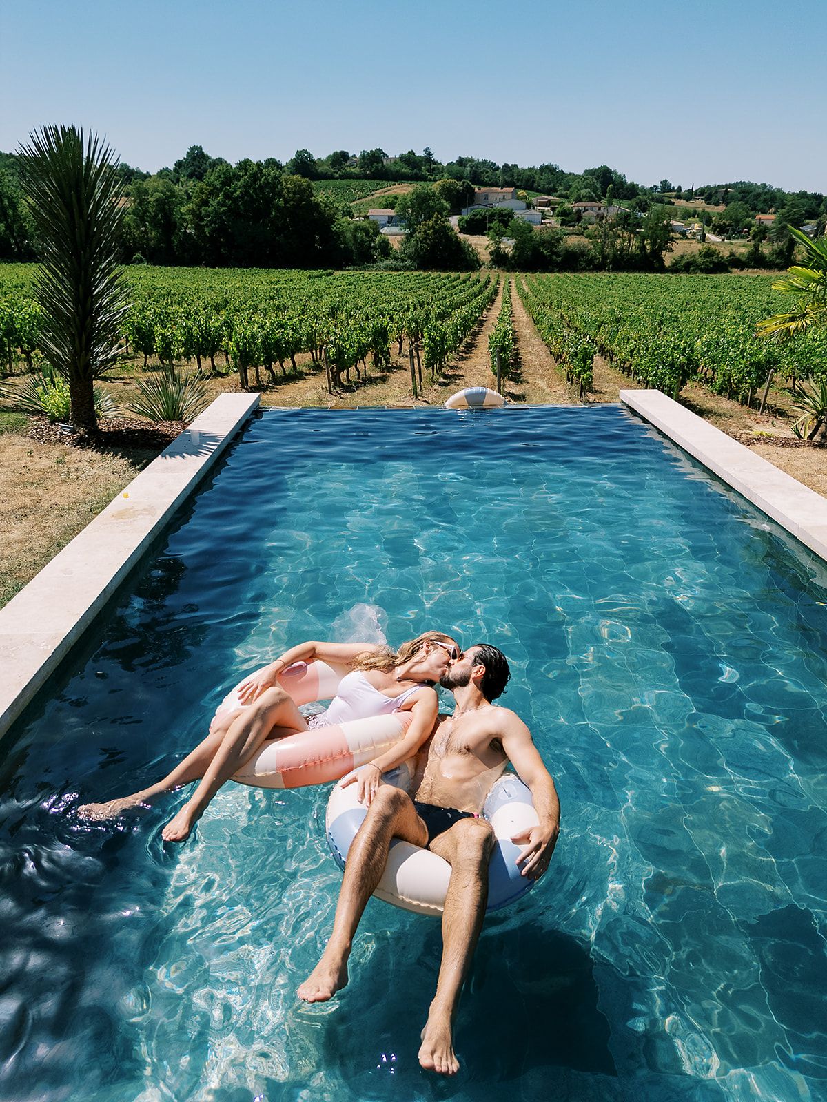 The couple lie together on a pale inflatable in a blue swimming pool, with vineyards stretching out behind them.