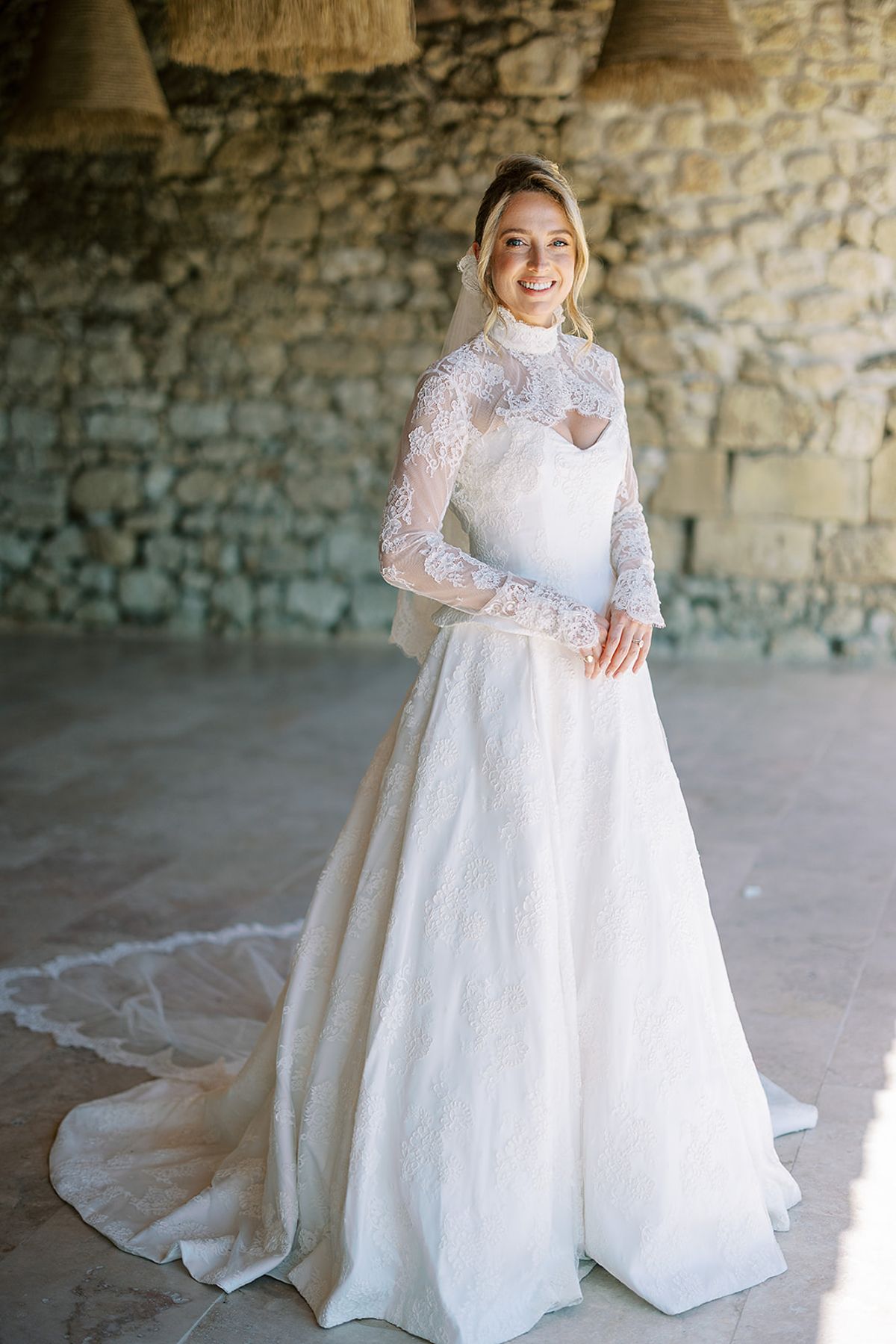 The bride stands smiling in a high-necked lace gown with a long train, in front of a rough stone wall.