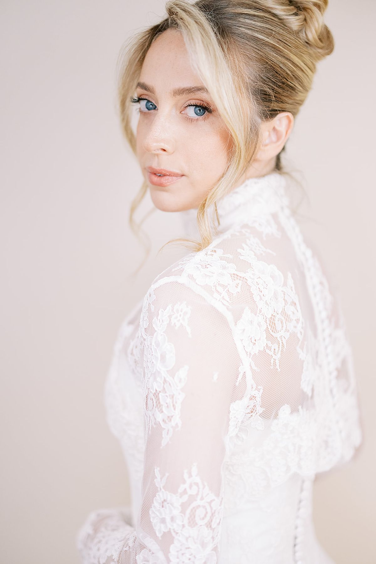 The bride, with blonde hair in an updo, looks over her shoulder at the camera, wearing a delicate lace high-necked gown.