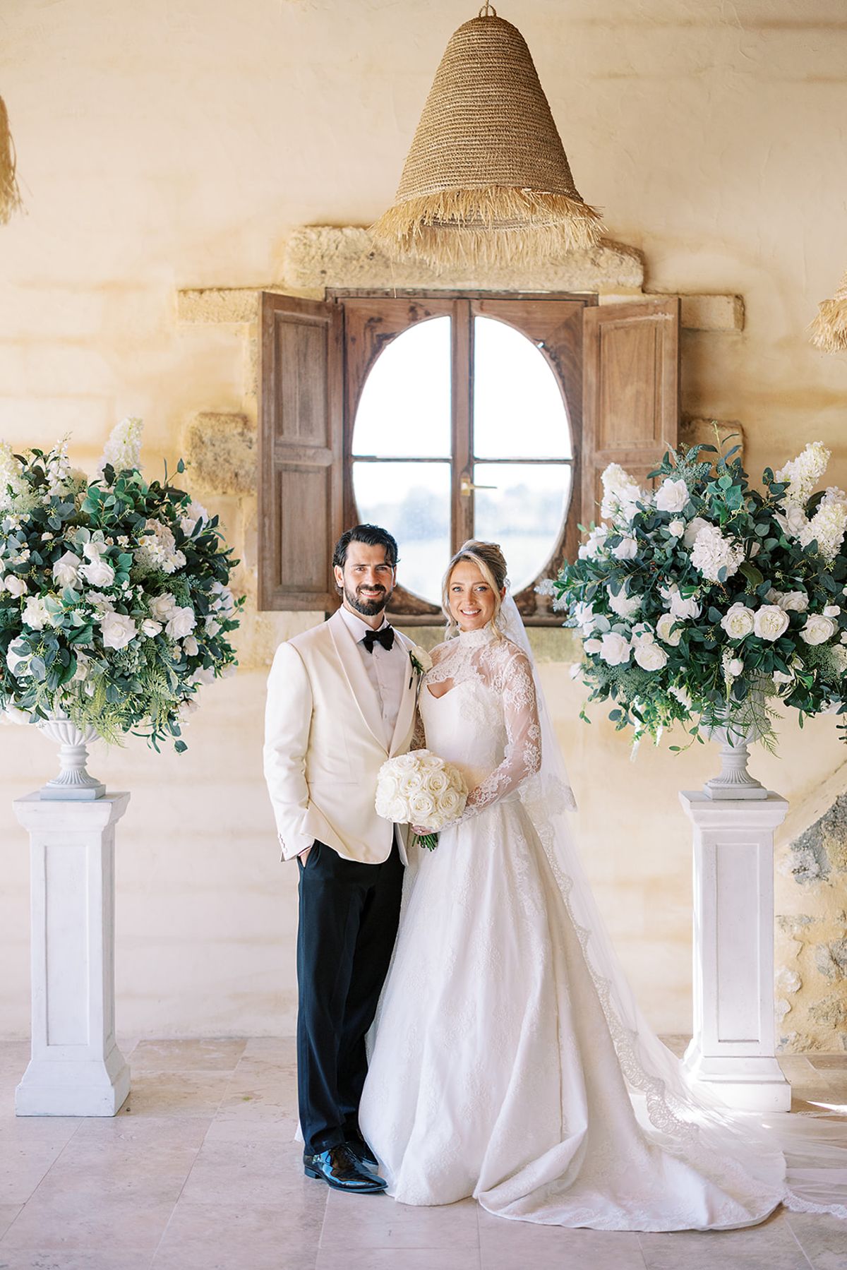 The couple stand together in front of an arched oval window, flanked by tall white flower arrangements on white pedestals.