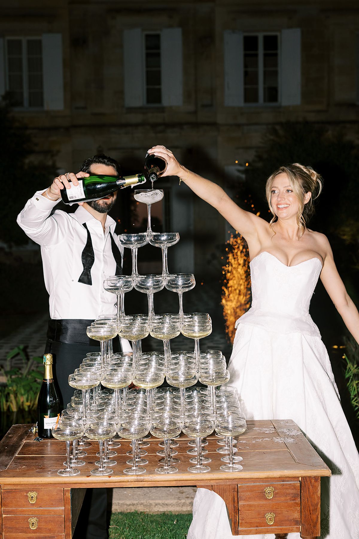 The couple pour champagne from two bottles over a tall pyramid of coupe glasses on a wooden table outside a stone building.