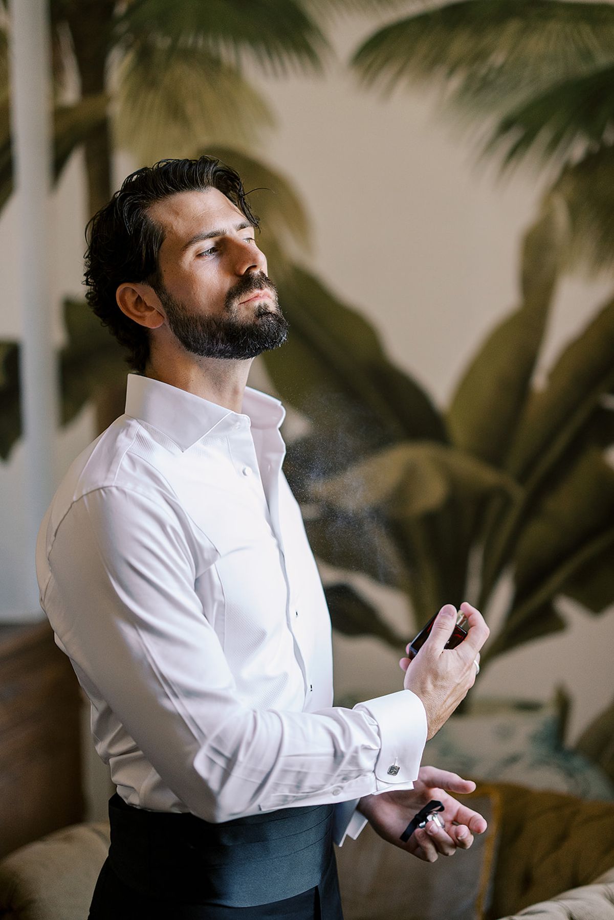 A bearded man in a white shirt looks upwards while fastening a cufflink in front of palm leaf wallpaper.