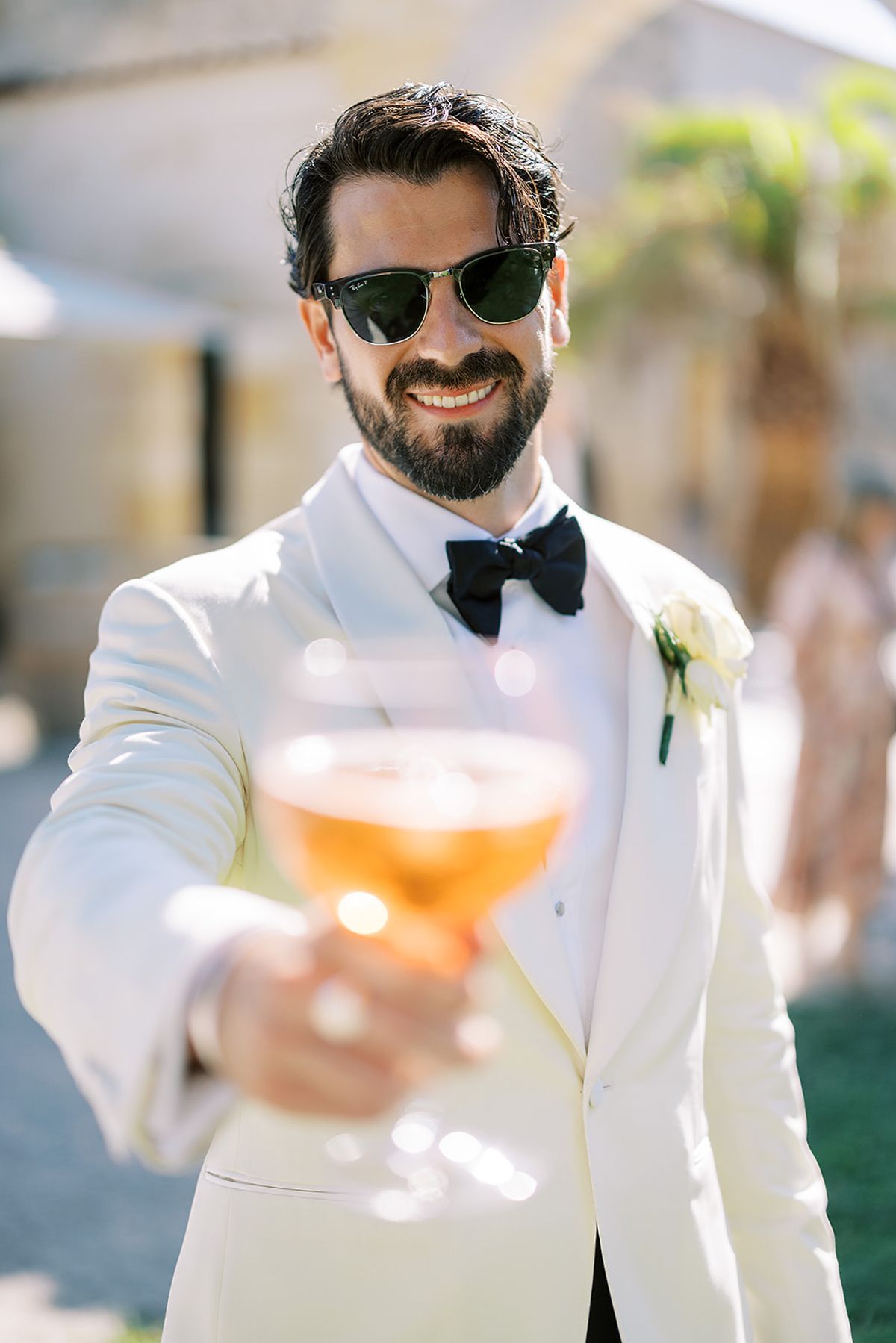 A smiling bearded groom in sunglasses and a white dinner jacket holds out a glass of amber drink towards the camera.