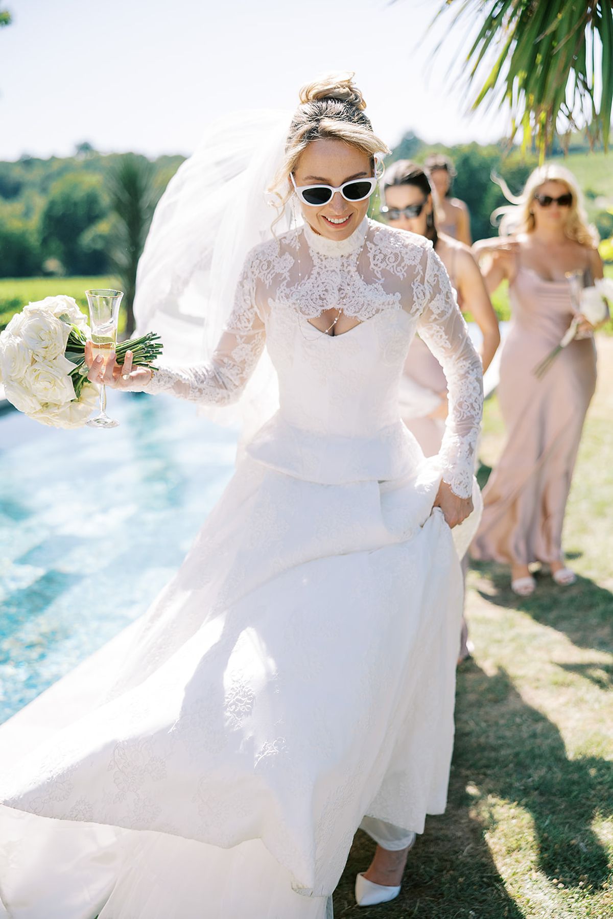 The bride in a lace gown and white sunglasses walks beside a pool, holding her skirt, with bridesmaids following behind.
