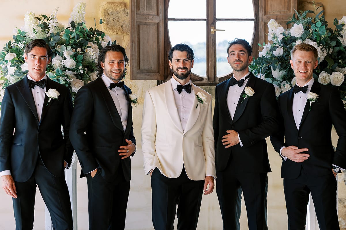 Five men in formal wear stand side by side, four in black dinner jackets and one in a cream jacket, before white flowers.