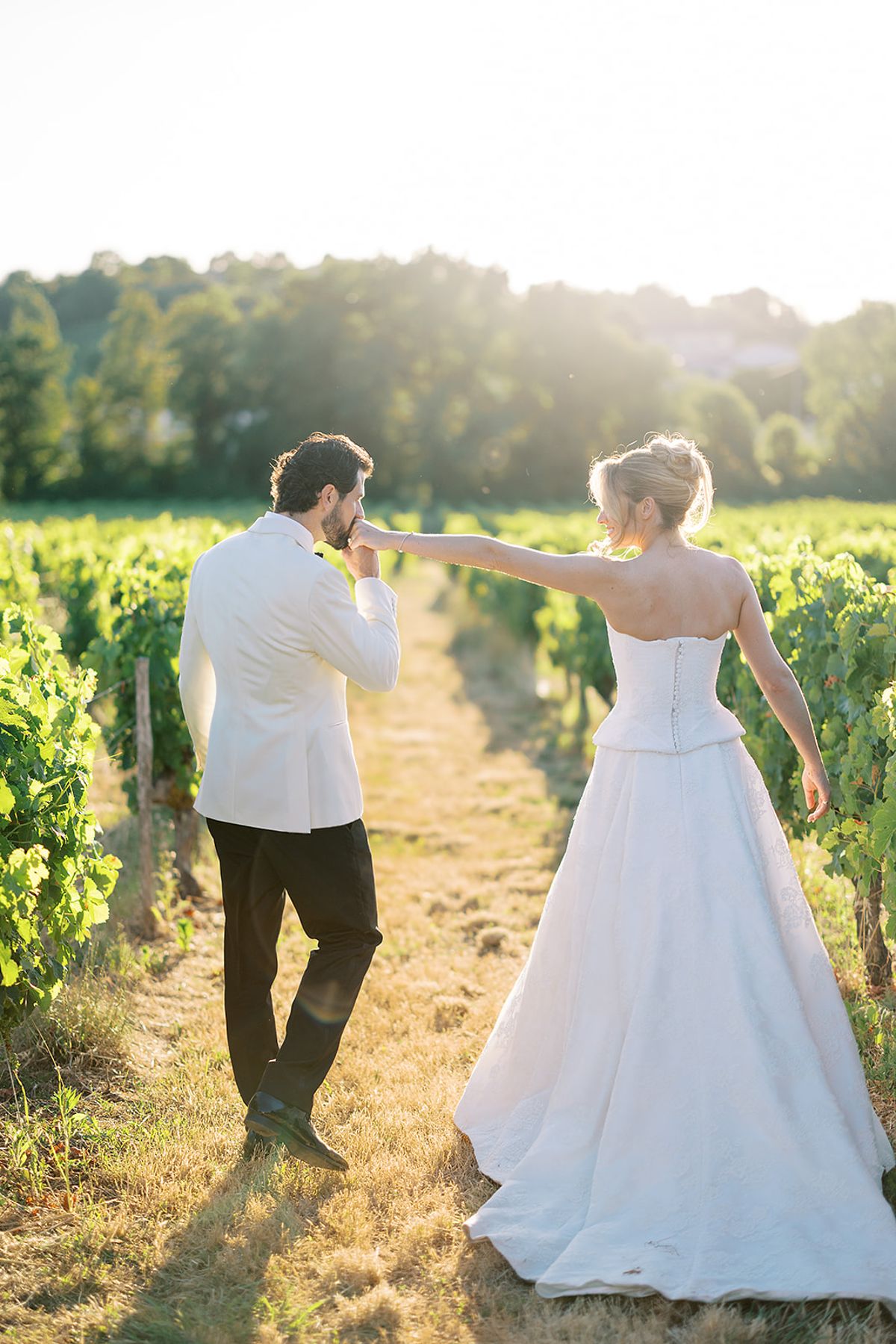 The groom in a white jacket kisses the bride's hand as they walk together along a grassy path between vines.