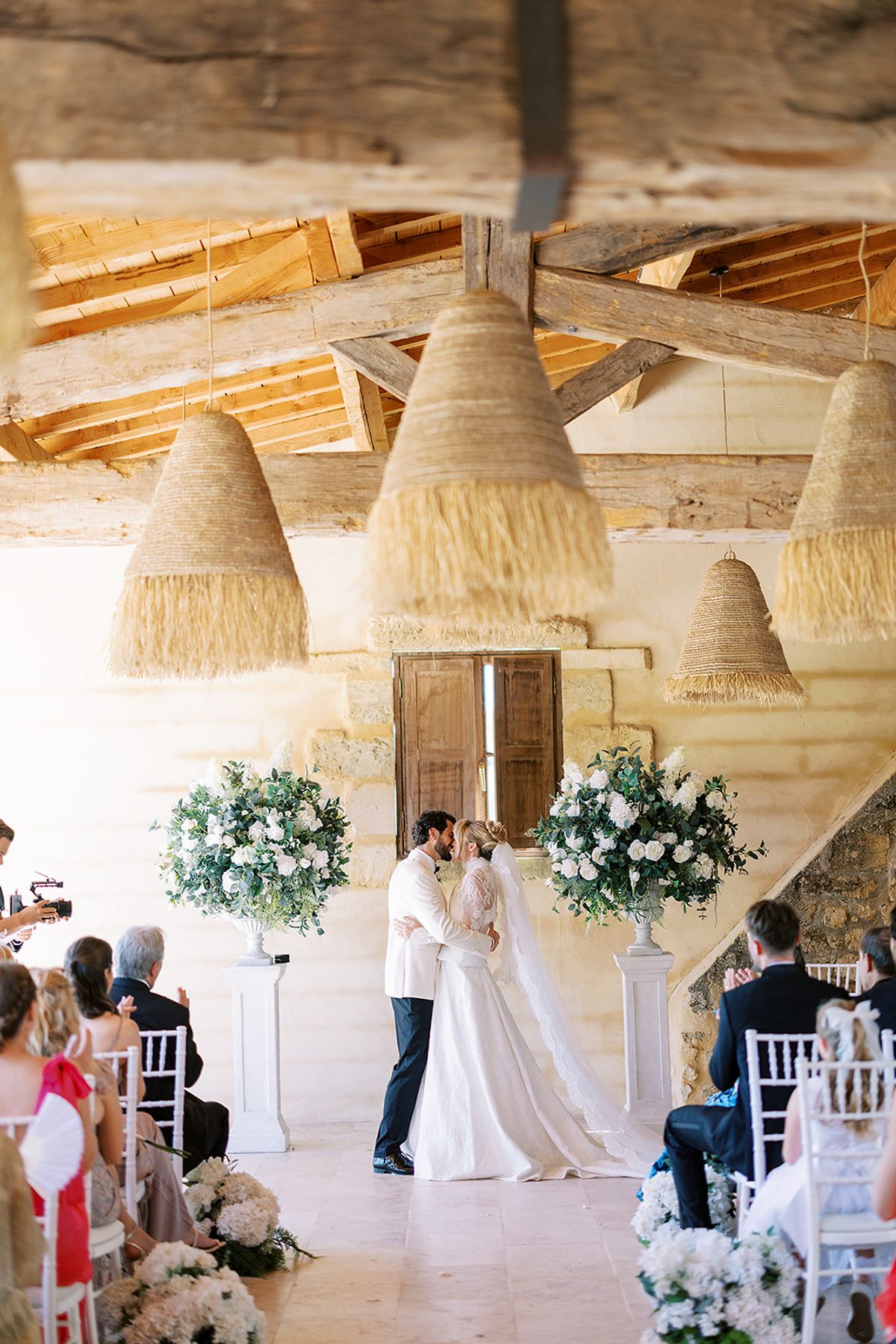The couple kiss between white flower arrangements as seated guests watch, under rustic beams and woven fringed lampshades.