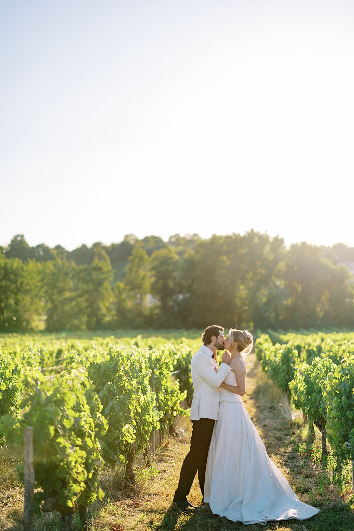 The couple share a kiss among rows of vines, with trees and a bright, hazy sky behind them.