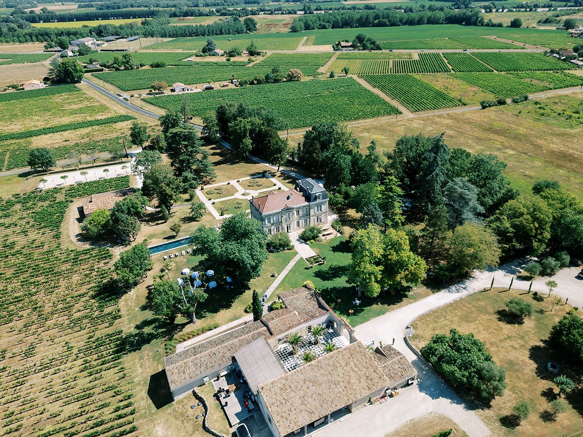 An aerial view shows a chateau and tiled outbuildings surrounded by trees, fields and rows of vines.