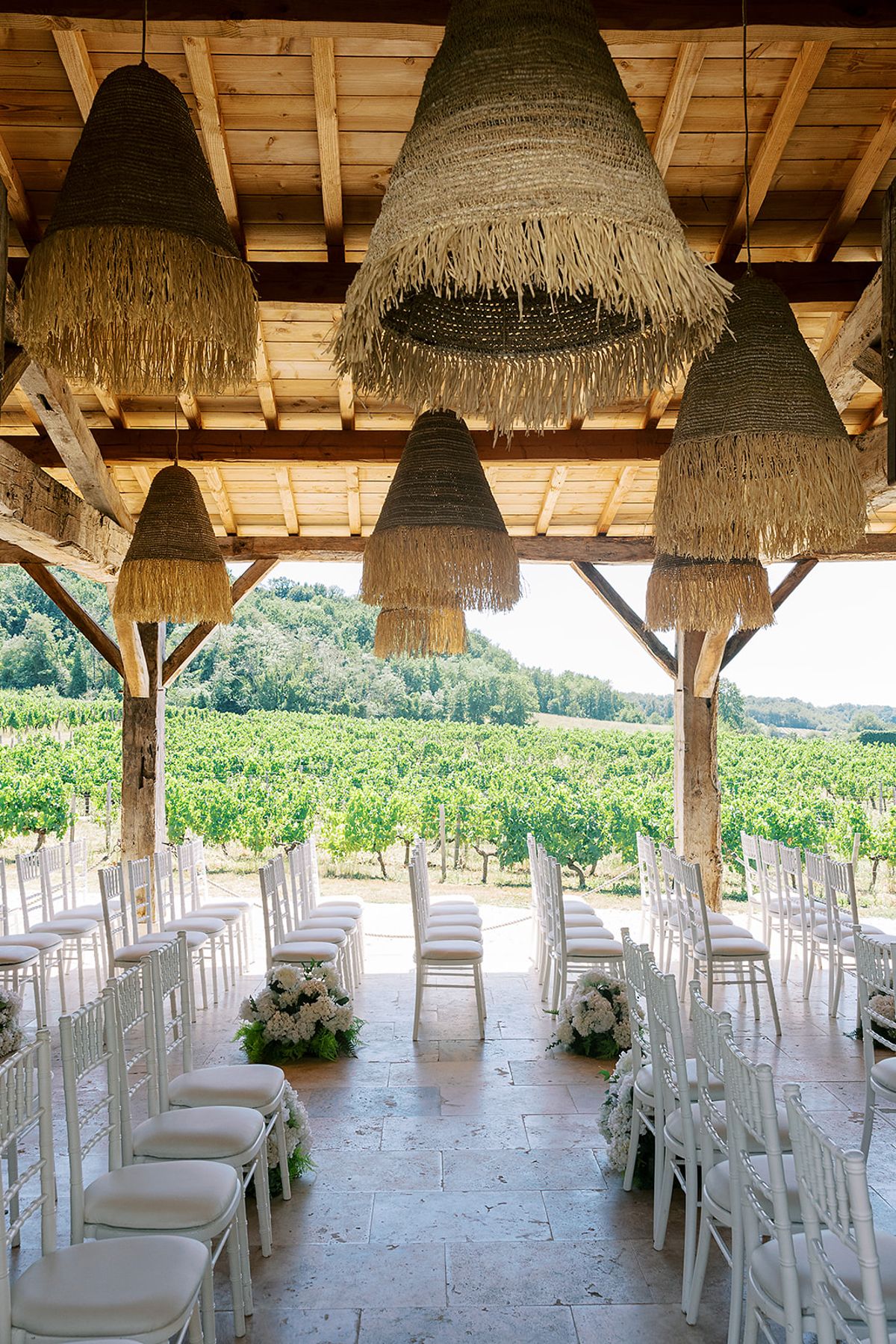 Rows of white chairs and white flower arrangements under a timber roof with raffia lampshades, facing a vineyard.