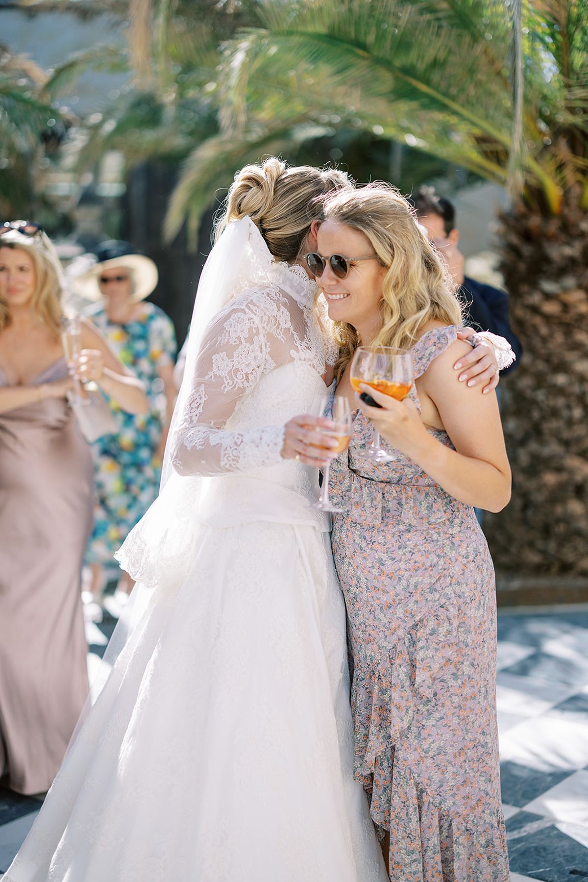 The bride in a lace veil hugs a woman in a floral dress, both holding drinks, with other guests behind them.
