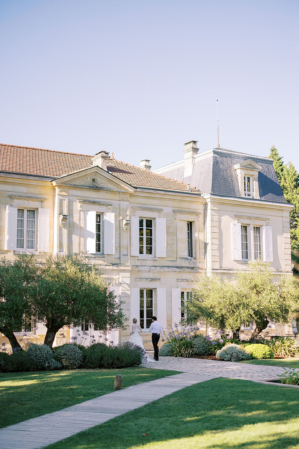 A couple in wedding attire walk towards a pale stone château with white shutters, olive trees and lawns in the sunshine.