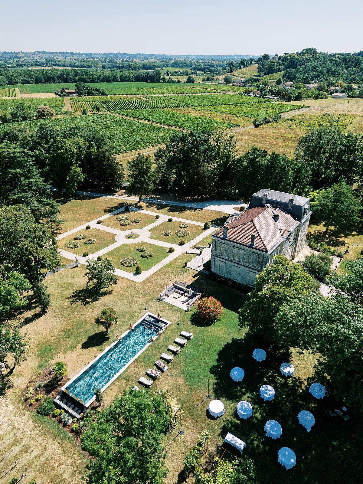 An aerial view of the château with its long pool, formal gardens and white parasols, surrounded by vineyards.