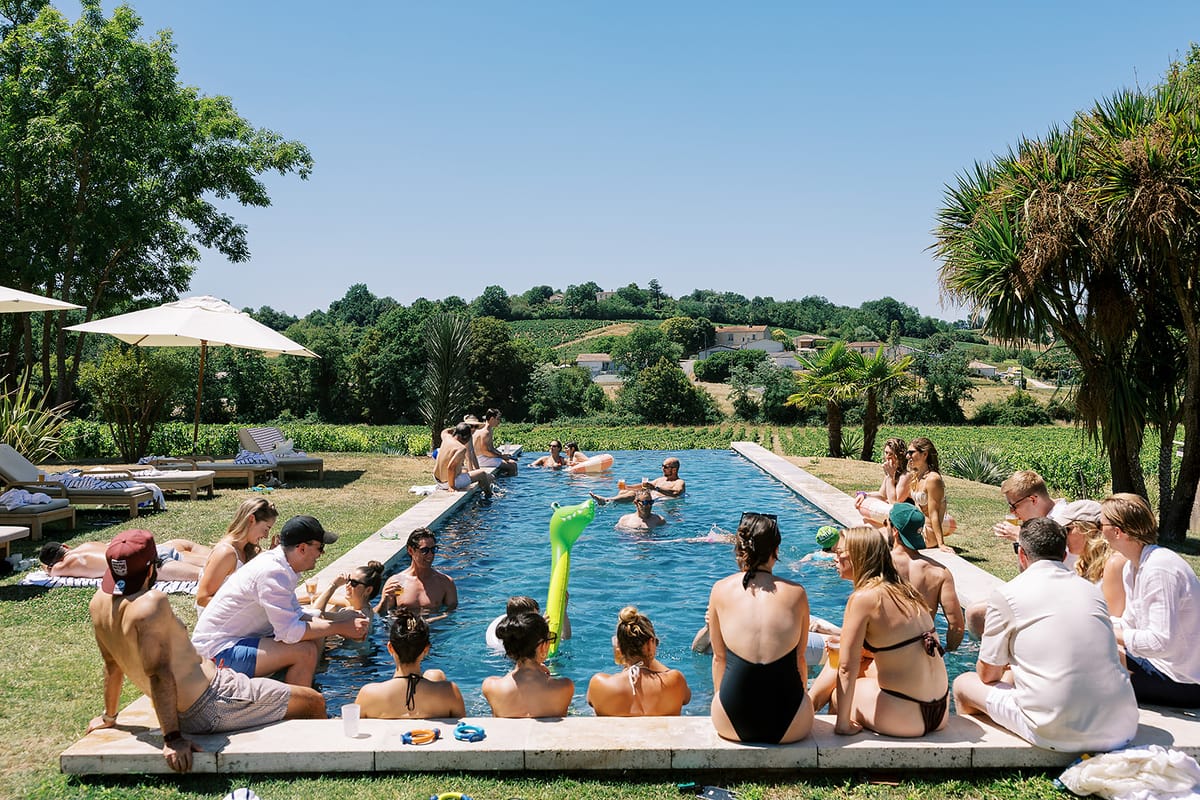 Guests sit along the edge of a long swimming pool and chat, with parasols, palm trees and vineyards behind them.
