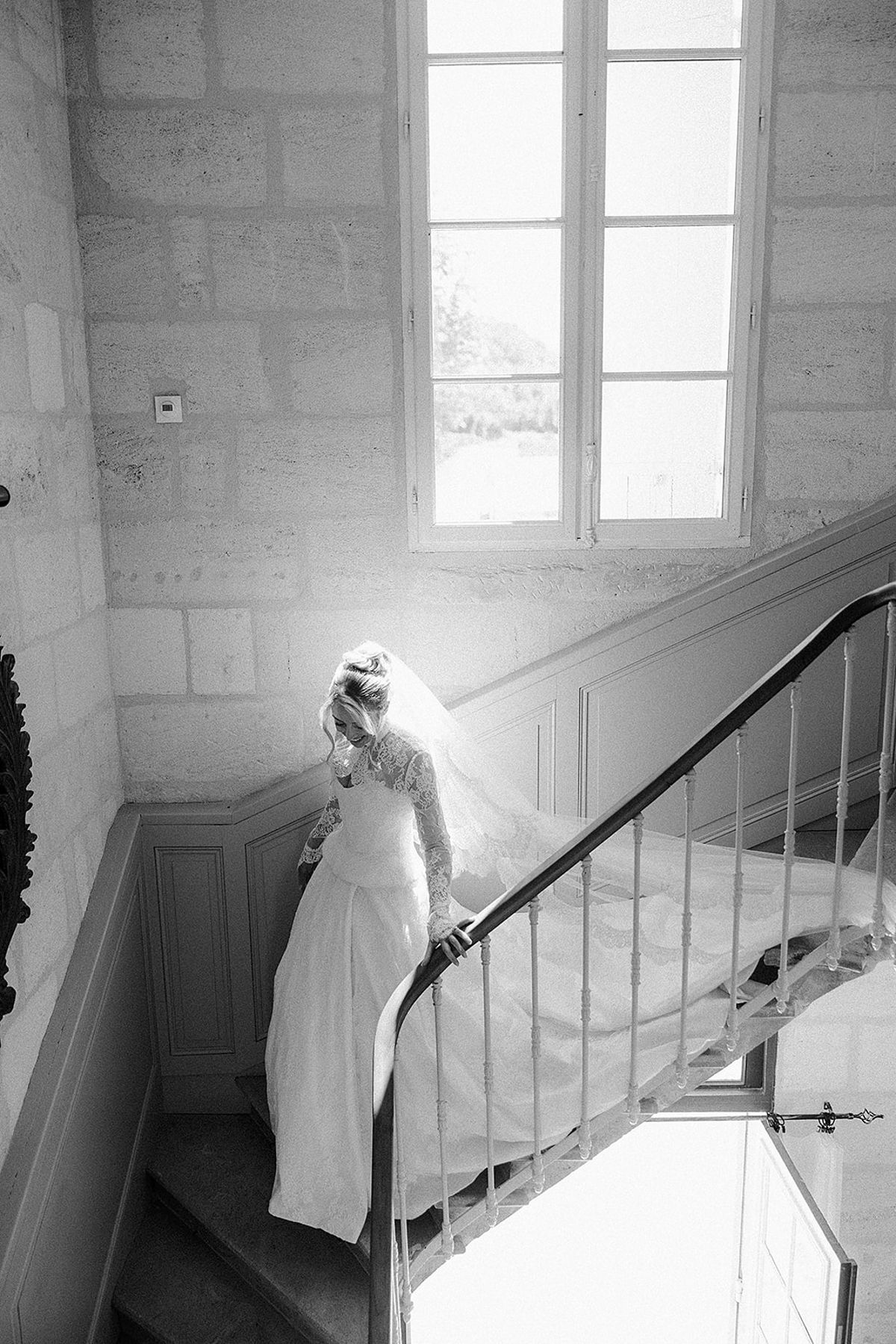 The bride in a lace-sleeved gown and long veil walks down a stone staircase, hand on the iron banister, in black and white.
