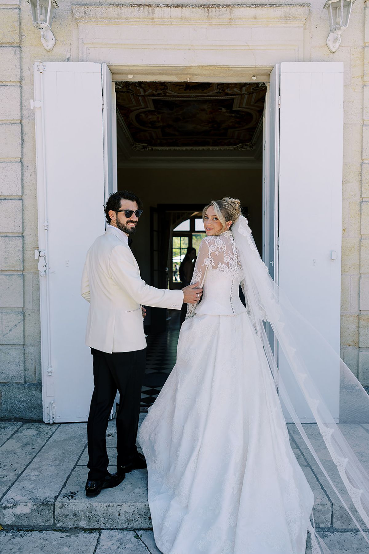 The couple, seen from behind, hold hands in a doorway with open white shutters, the bride's long veil and train trailing on the stone.