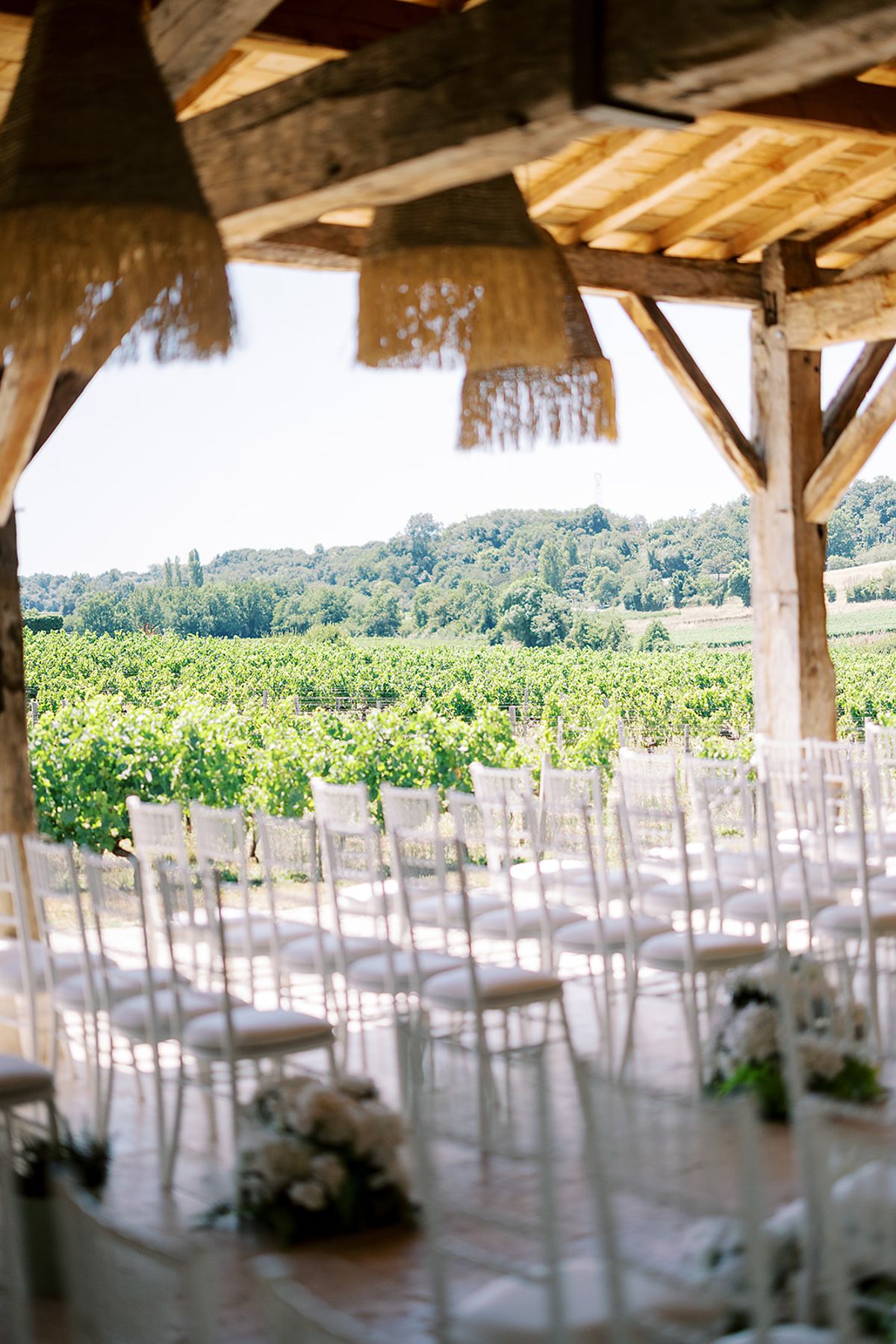 Rows of white chairs sit in a timber pavilion with a thatched edge, looking out over green vineyards.