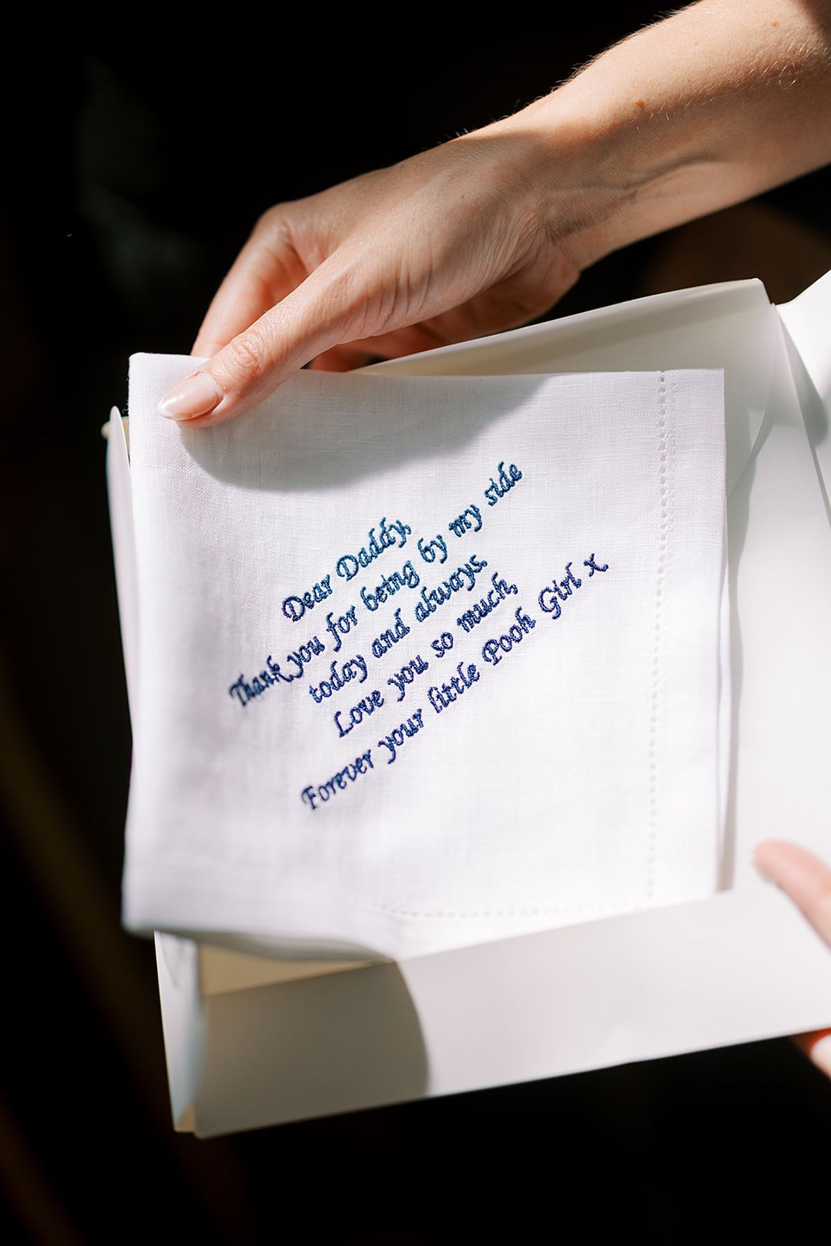A hand holds a white embroidered handkerchief in a box, stitched in blue with a thank-you message to "Daddy".