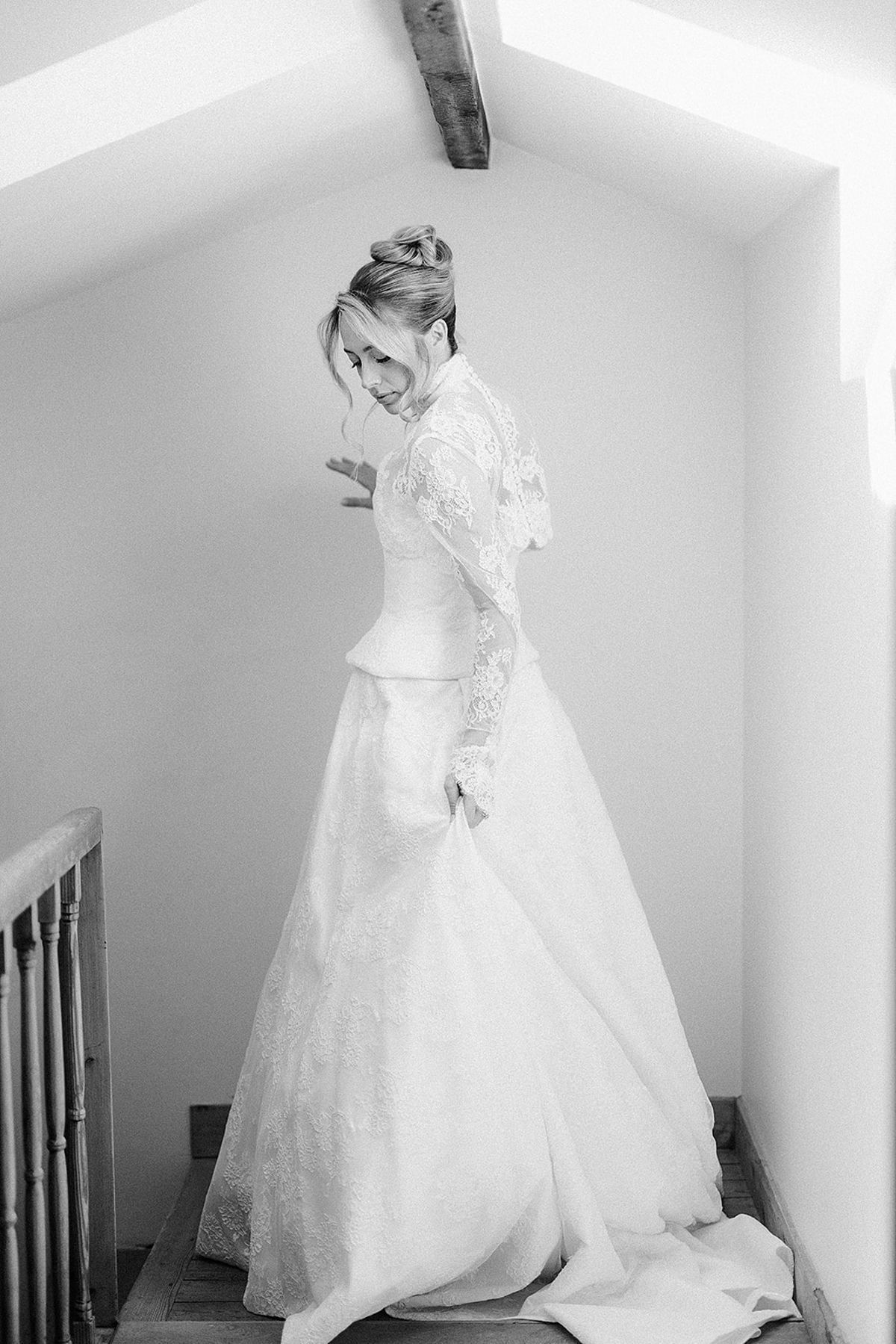 In black and white, the bride in a lace-sleeved ball gown stands at the top of a staircase, looking down.