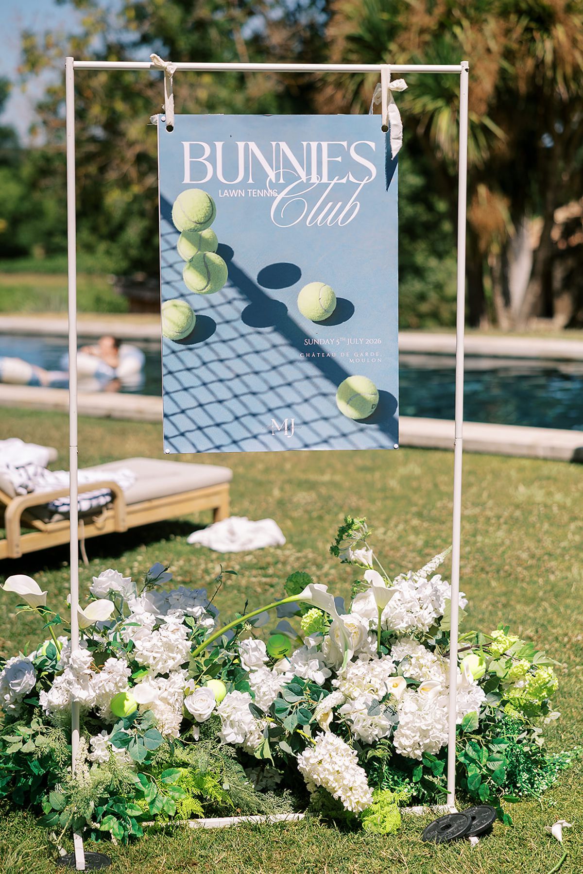A tennis-themed welcome sign hangs on a white frame above white flowers on the lawn beside the pool.
