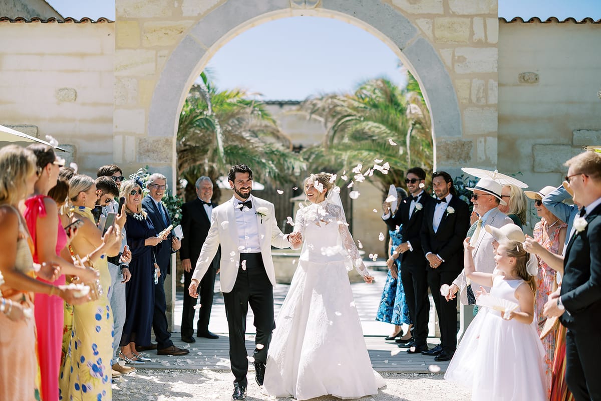 The couple walk through a line of smiling guests in a sunlit courtyard, framed by a stone archway and palm trees.