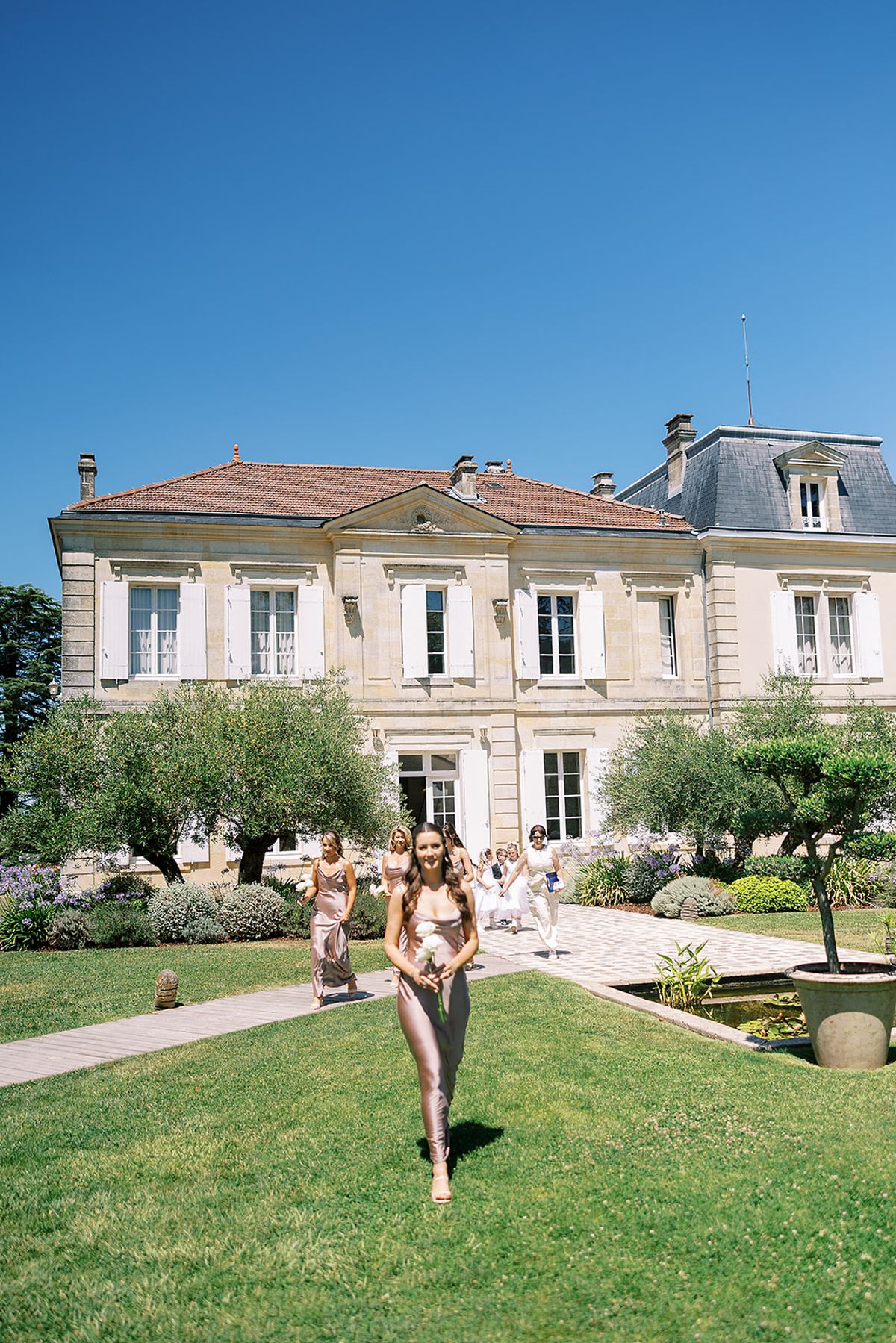 A woman in a satin blush dress walks across a lawn, with other guests behind her, before a cream manor house.