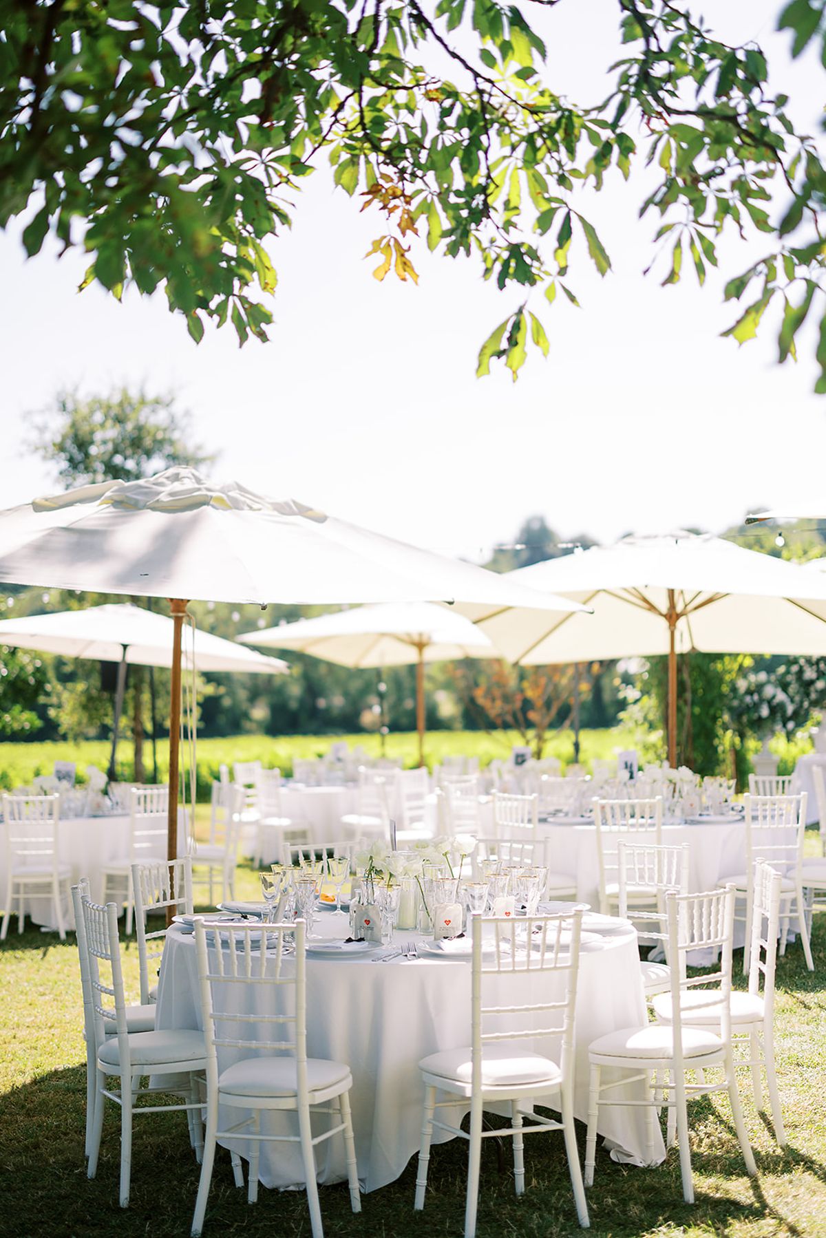 Round tables with white cloths and white chairs are set on a lawn beneath cream parasols and tree branches.