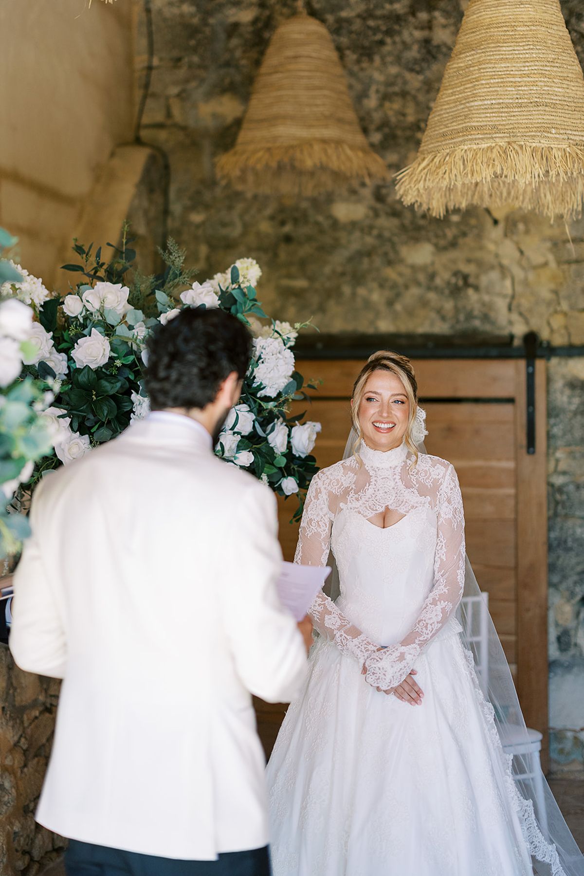 The groom, seen from behind, faces the bride in a high-necked lace gown in front of white flowers and a barn door.