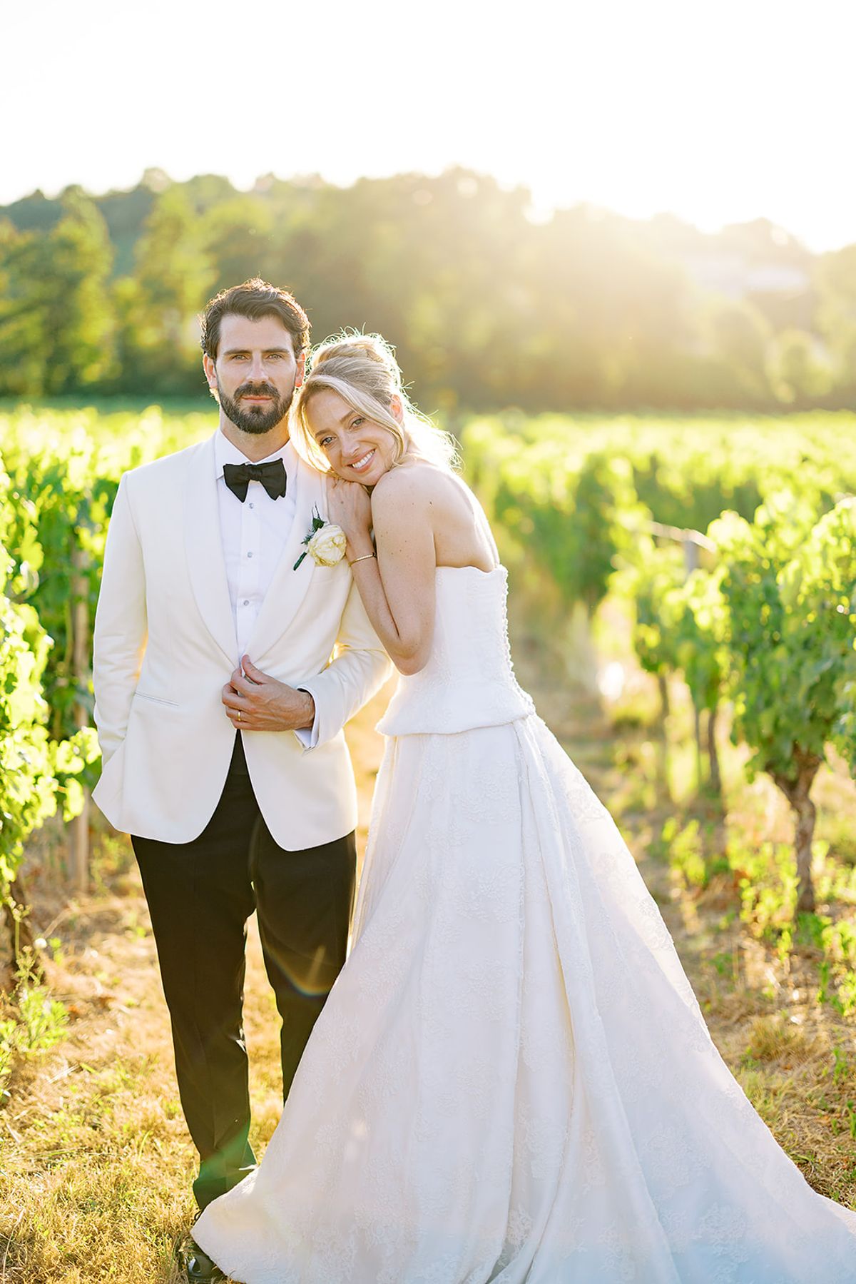 The couple stand close together among rows of vines, the groom in a white dinner jacket and the bride in a white gown, smiling at the camera.