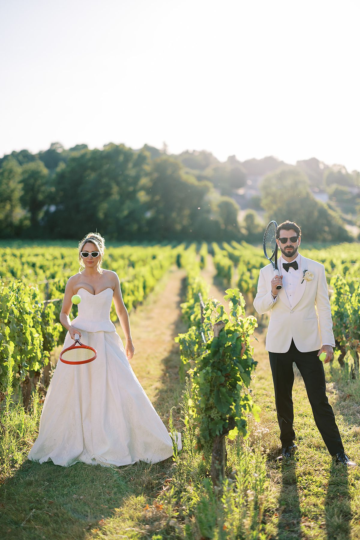 The bride, holding a red hoop, and the groom in a white jacket stand apart between rows of vines, both wearing sunglasses.