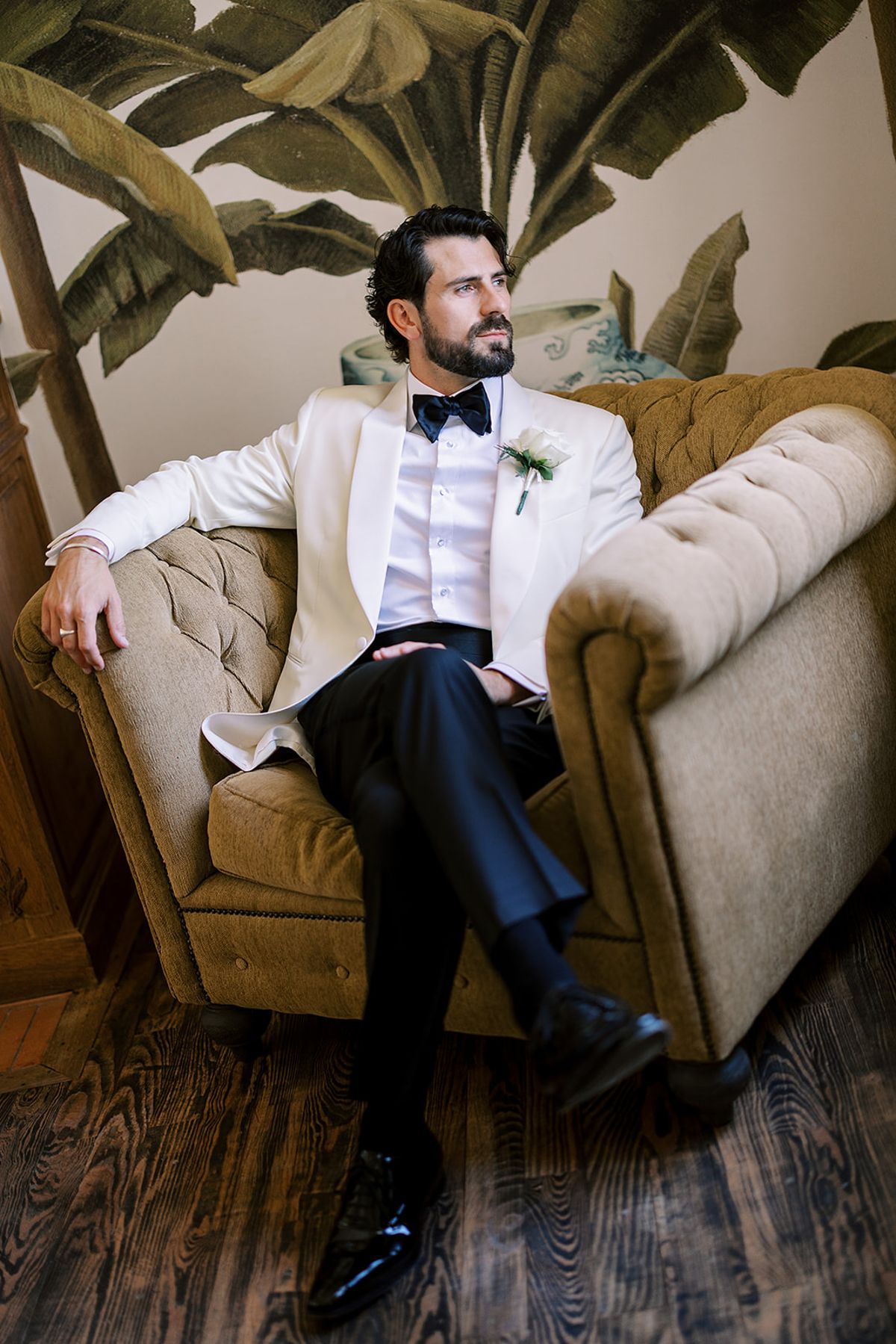 The groom in a white dinner jacket and bow tie sits with crossed legs in a tufted armchair before banana-leaf wallpaper.
