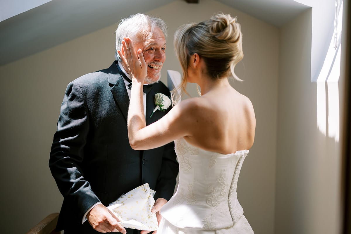 The bride in a corset-back gown gently touches the cheek of a smiling older man in a dark suit.