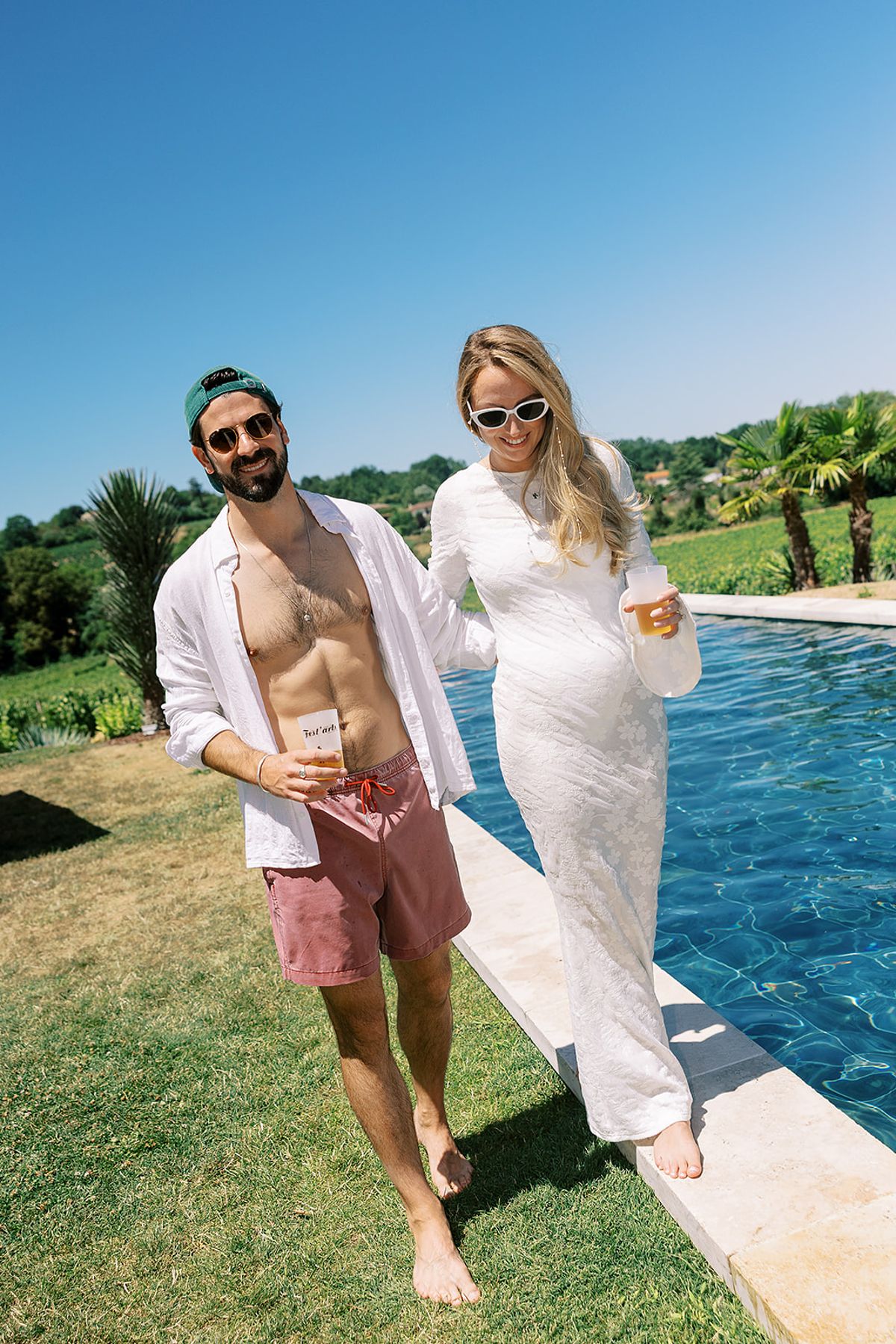 The groom in an open shirt and shorts and the bride in a lace dress stroll barefoot beside a bright blue pool.