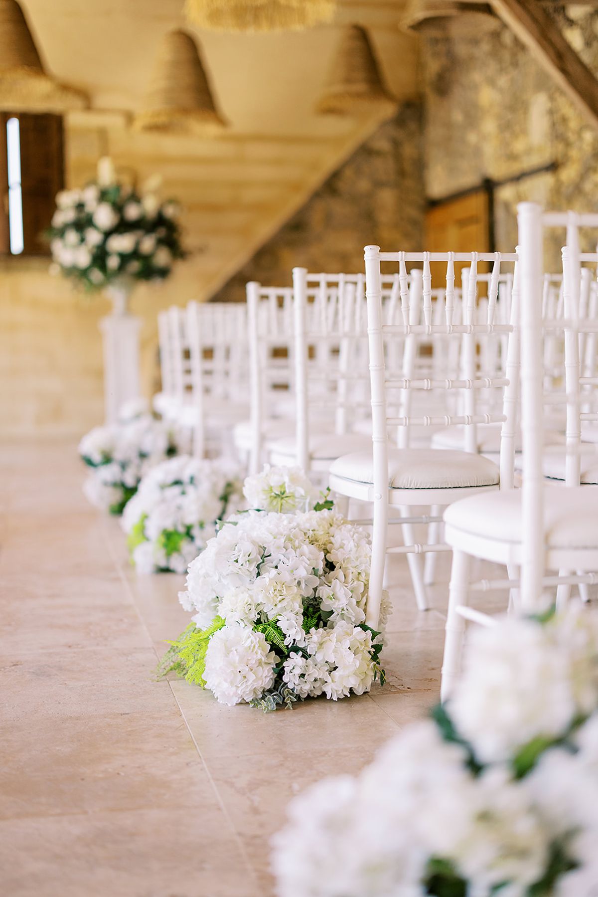 Rows of white chairs line an aisle decorated with clusters of white flowers beneath a rustic ceiling hung with woven lampshades.