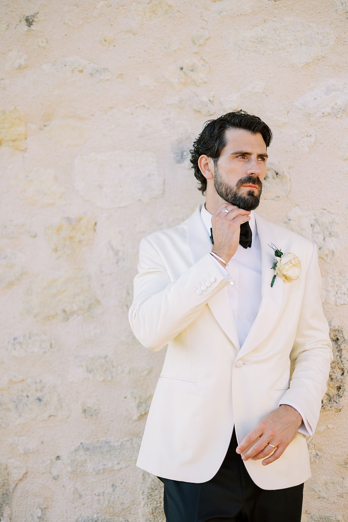 The groom in a white dinner jacket stands against a pale stone wall, touching his bow tie and looking away.