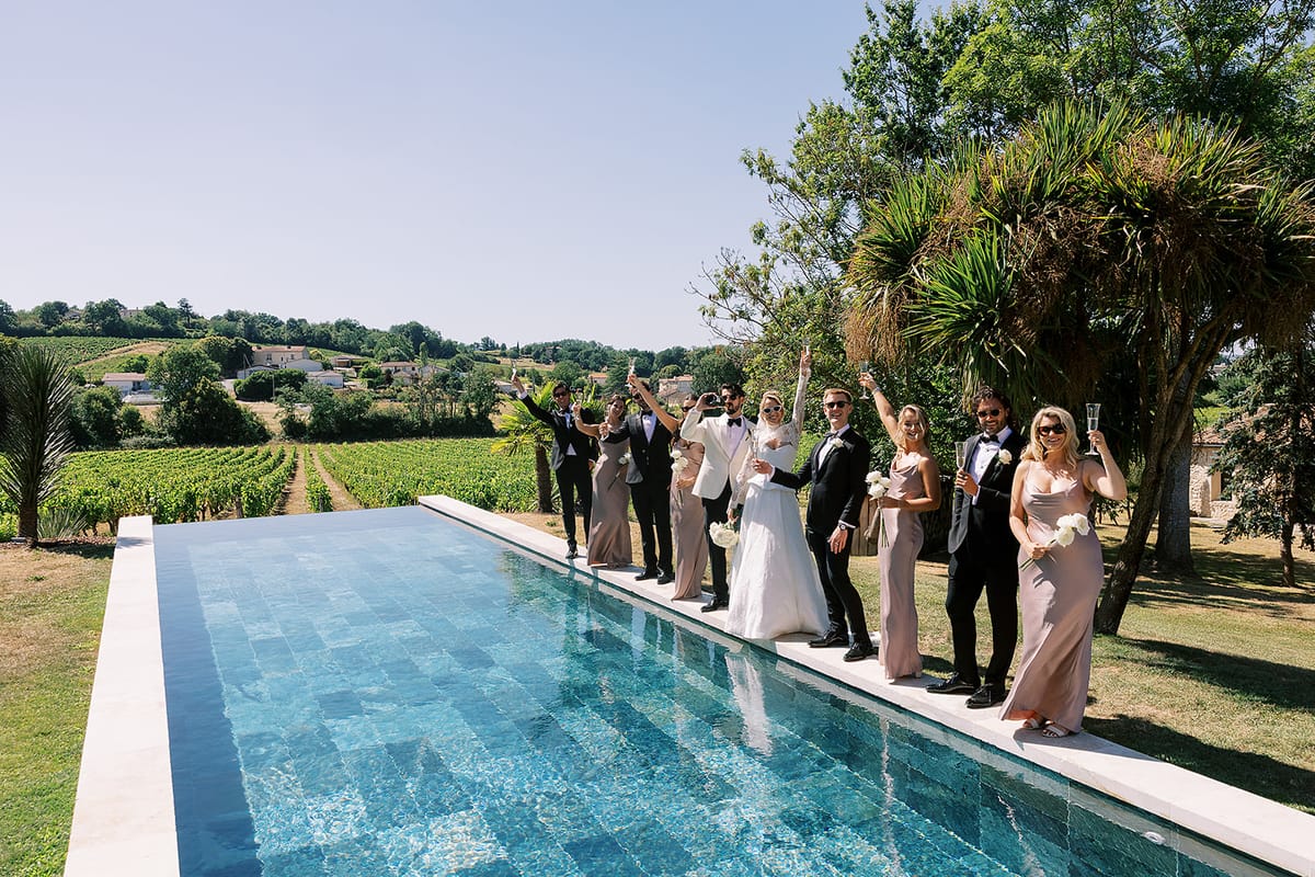 A group of wedding guests in formal clothes stand along the edge of a long turquoise swimming pool, with vineyards and trees beyond.