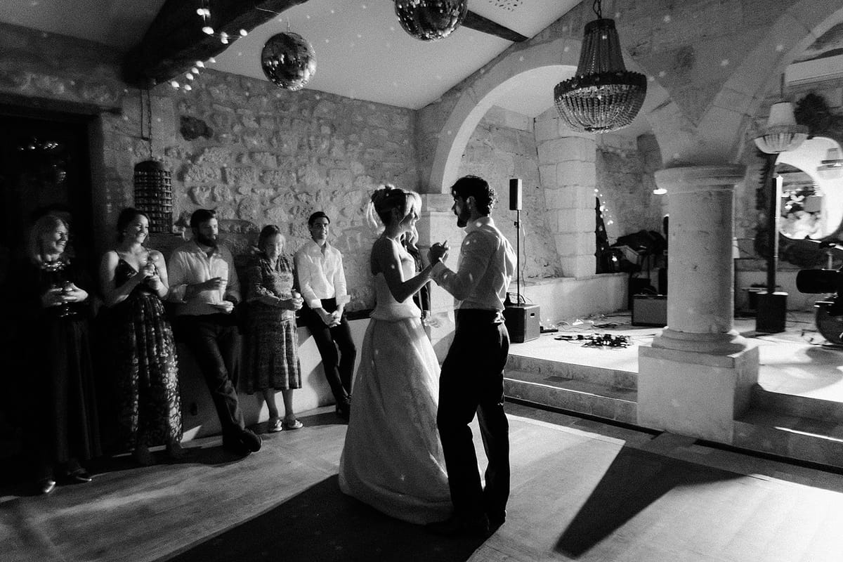 In black and white, the couple dance in a stone-walled room under a chandelier and disco ball while guests look on.