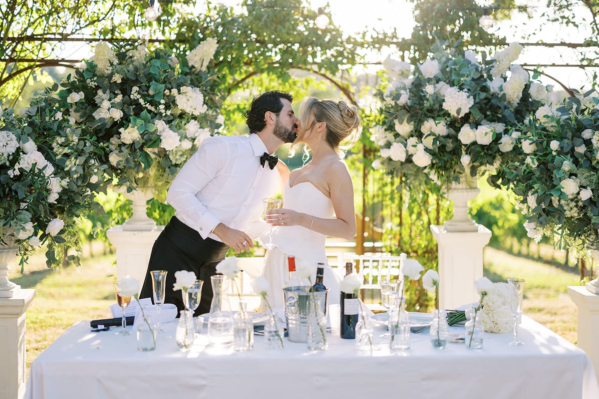 The couple kiss behind a white-clothed table set with glasses and bottles, framed by large white floral arrangements.