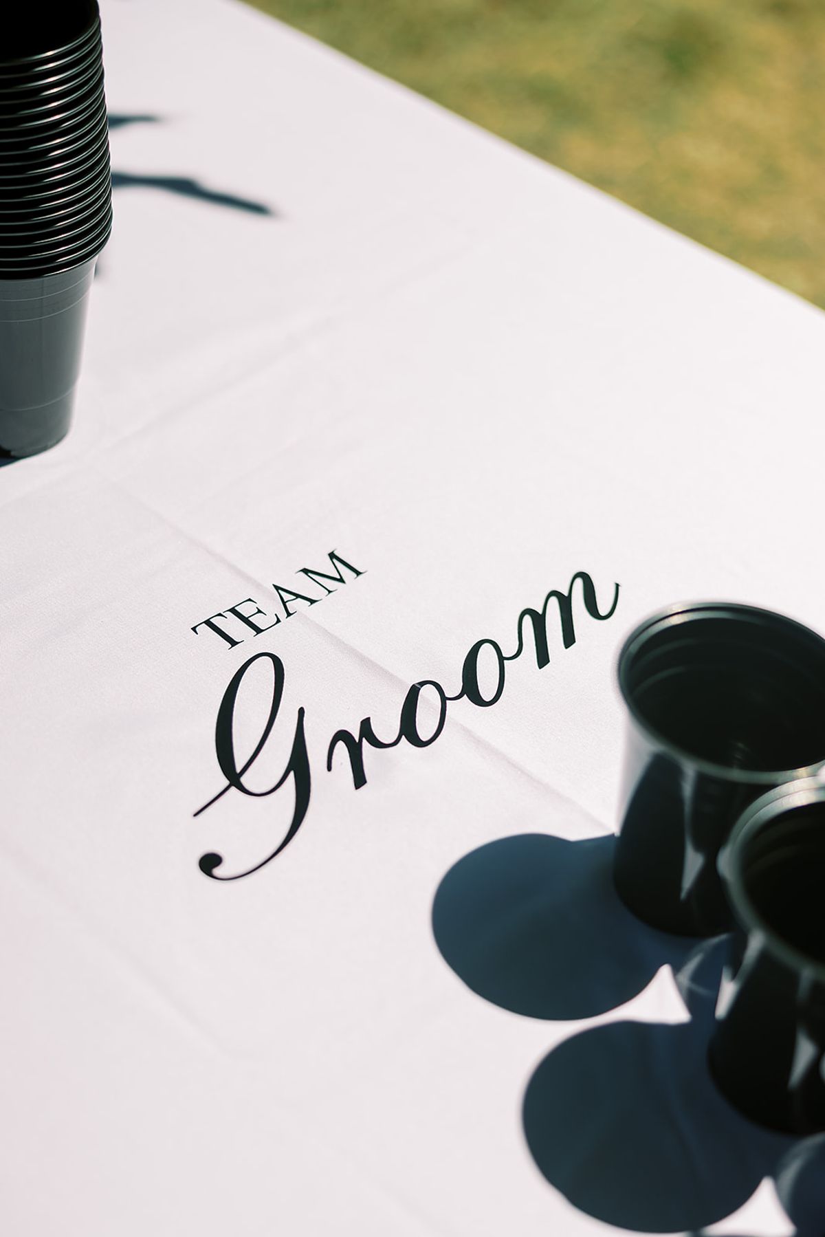 A white cloth printed with the words Team Groom in black script lies beside dark cups casting shadows.