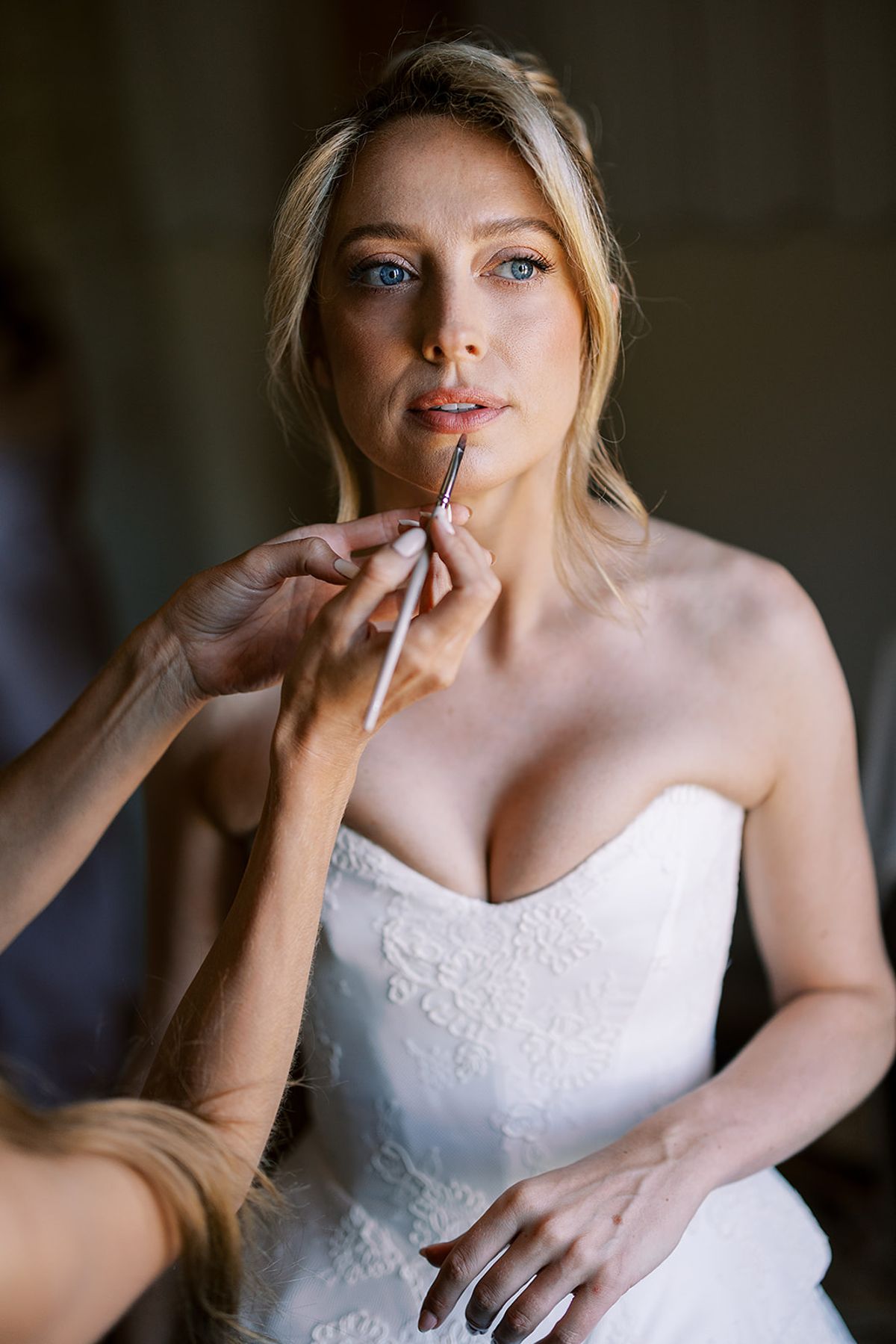 The bride in a strapless white lace gown has lipstick applied with a small brush by someone whose hands are visible.