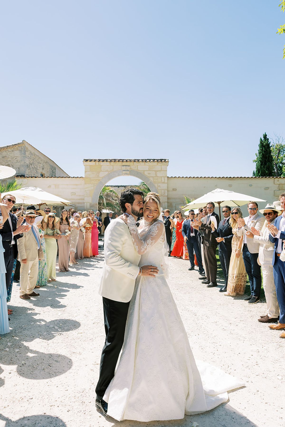 The couple kiss on a gravel path lined with applauding guests, with a stone archway and clear blue sky behind them.