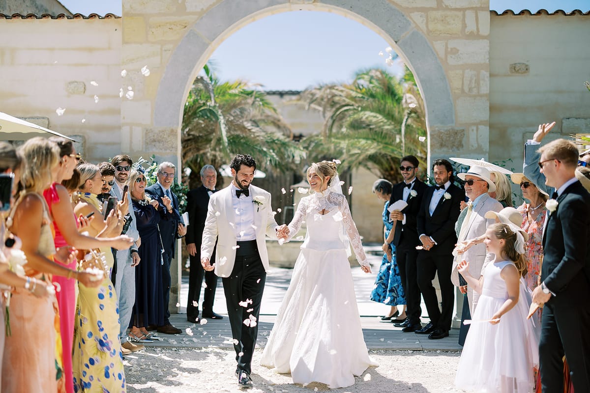 The couple walk hand in hand through a gravel courtyard as colourfully dressed guests line the path and throw confetti.